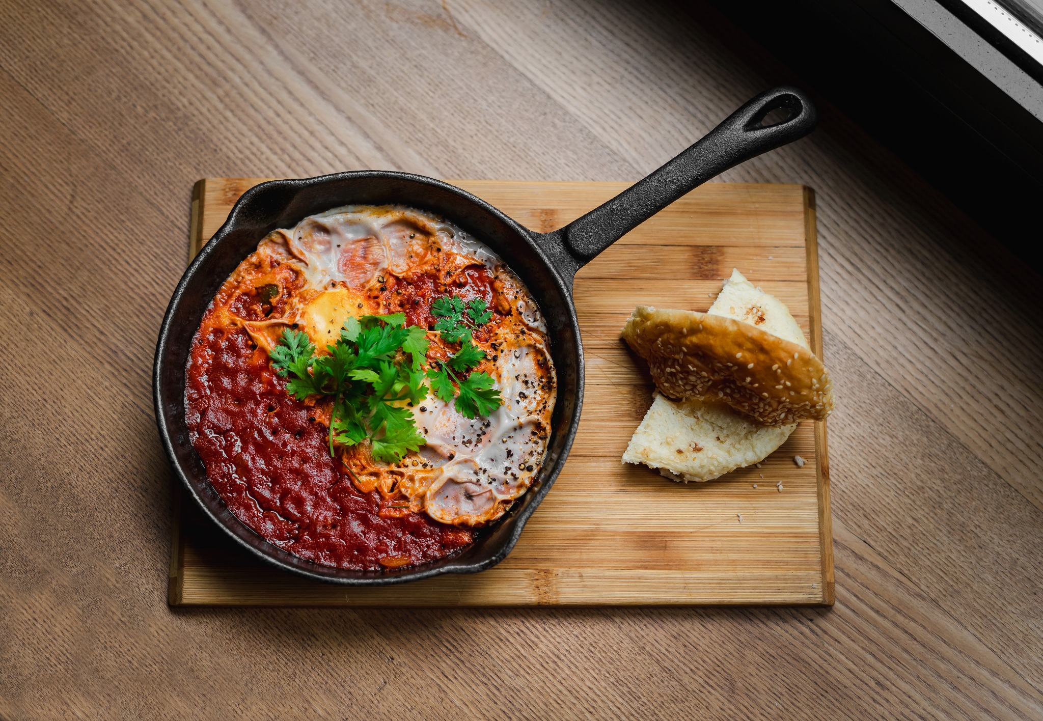 Shakshuka w żeliwnej patelni, udekorowana świeżą natką pietruszki, podana z chlebem na desce do krojenia, widok z góry.