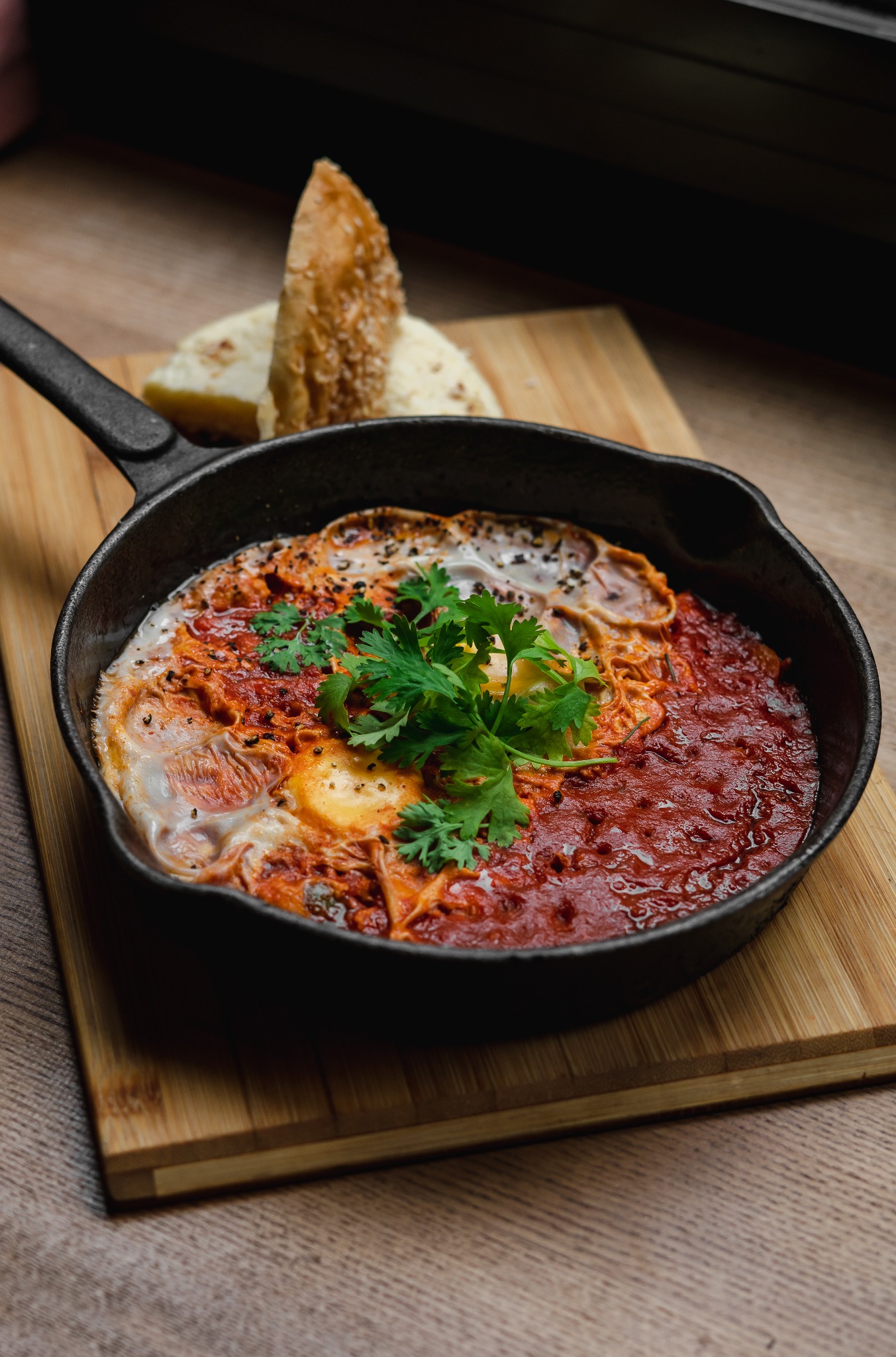 Shakshuka z jajkami sadzonymi w żeliwnej patelni, udekorowana świeżą kolendrą, podana z pieczywem na drewnianej desce.