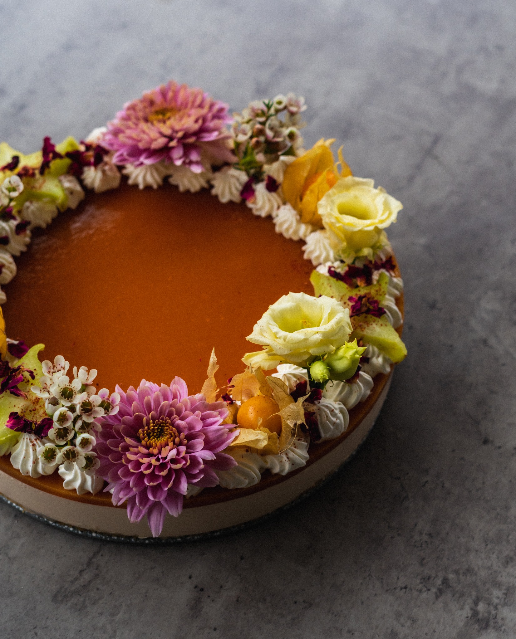 Kremowy tort z gładką, pomarańczową polewą, ozdobiony świeżymi kwiatami, bezikami i physalis na szarym tle.