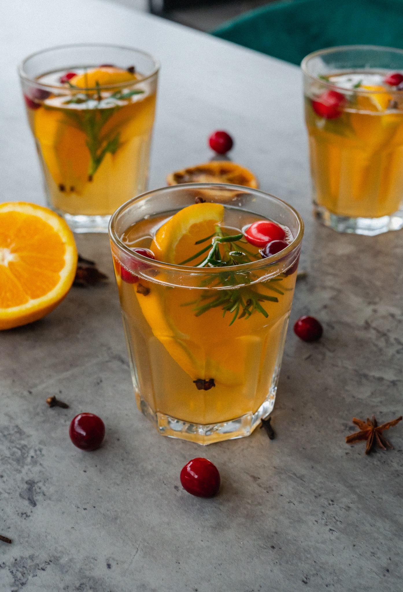 Trzy szklanki z napojem pomarańczowo-żurawinowym, ozdobione rozmarynem i goździkami, na szarym tle z plasterkiem pomarańczy i anyżem gwiaździstym.