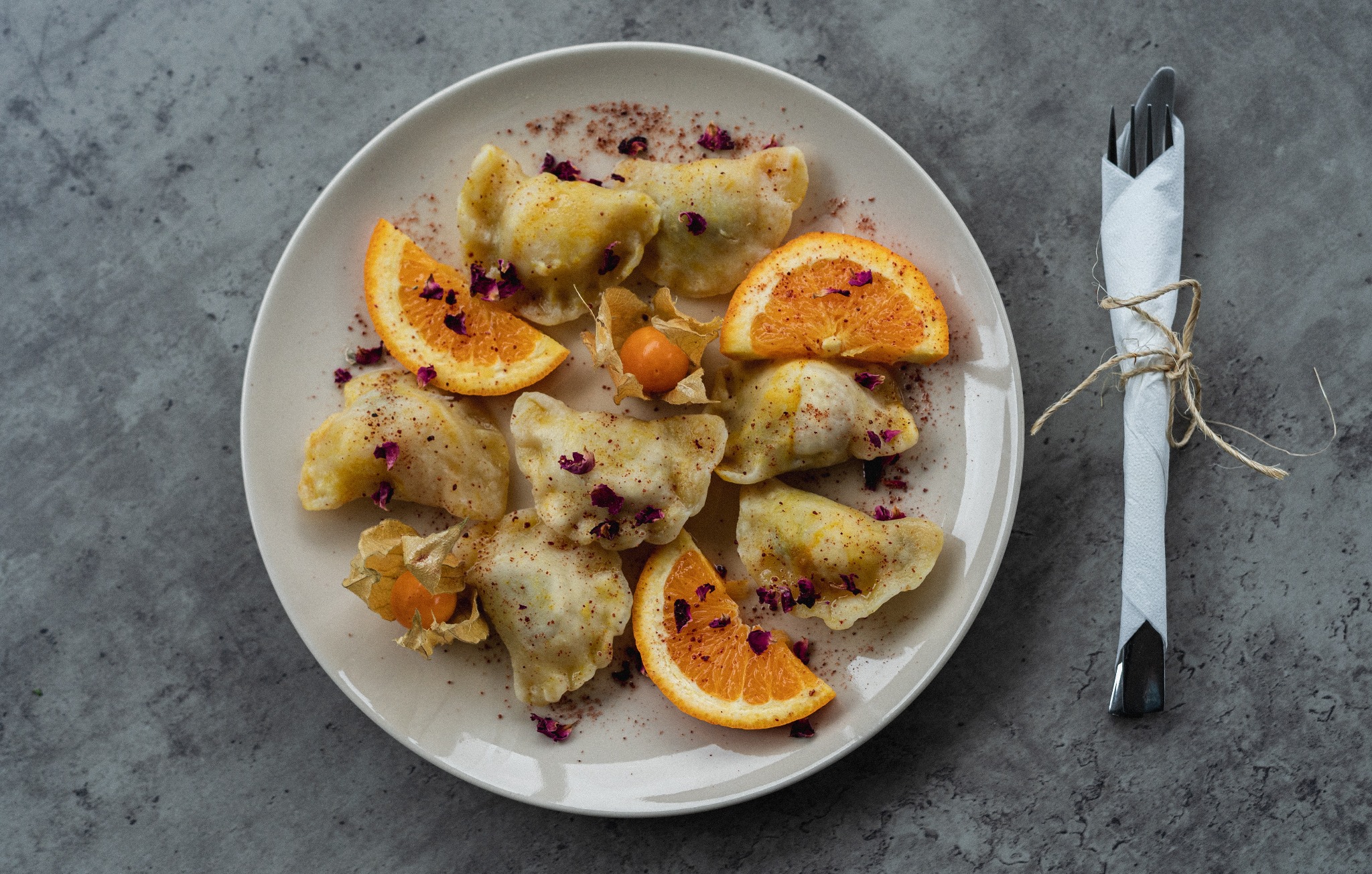 Kompozycja z pierogów, pomarańczy i miechunki na talerzu, posypana suszonymi płatkami róż, obok sztućce zawiązane sznurkiem.