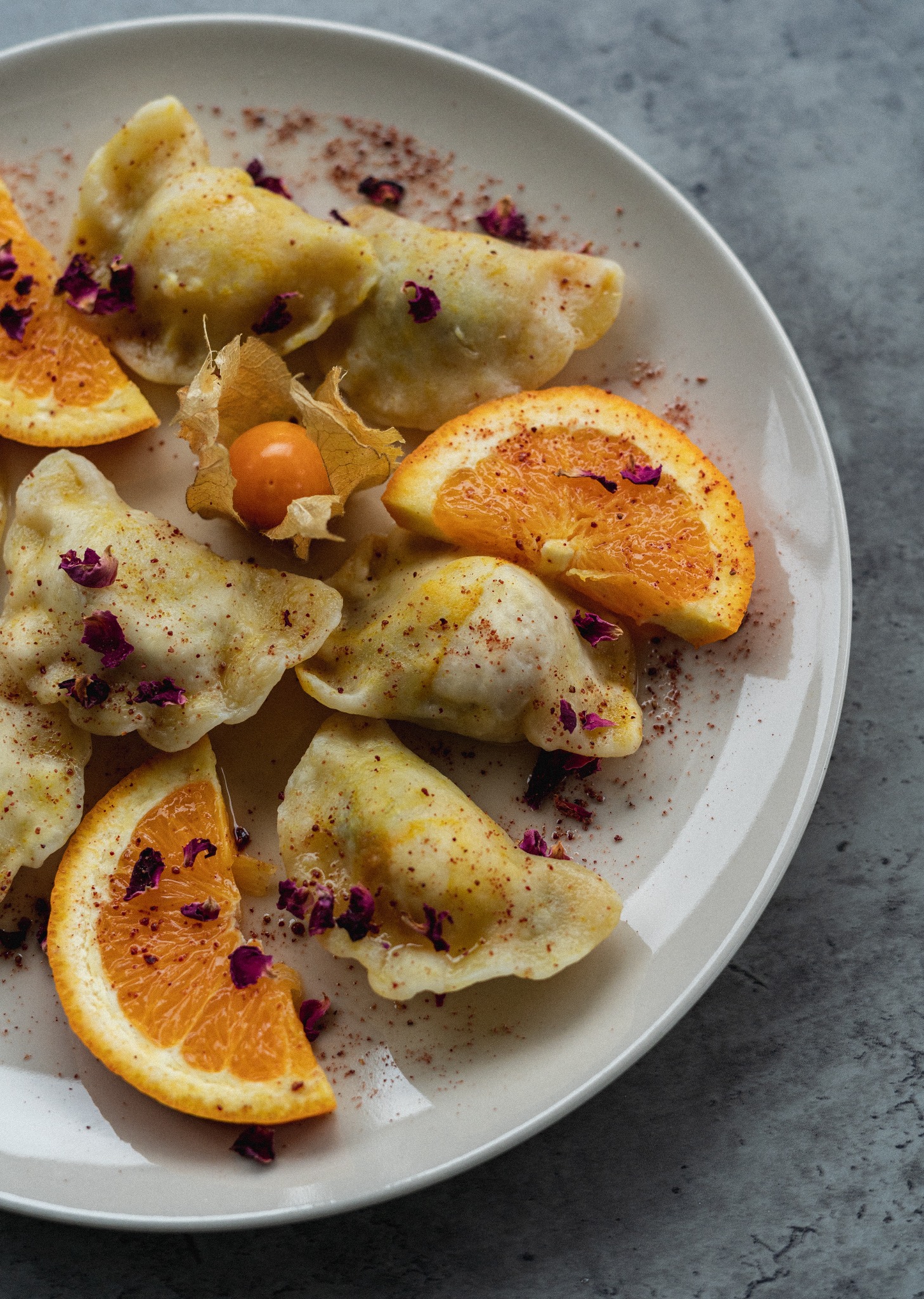 Elegancko podane pierogi z owocami: plasterki pomarańczy i miechunka peruwiańska, posypane suszonymi płatkami róż i przyprawami na białym talerzu.