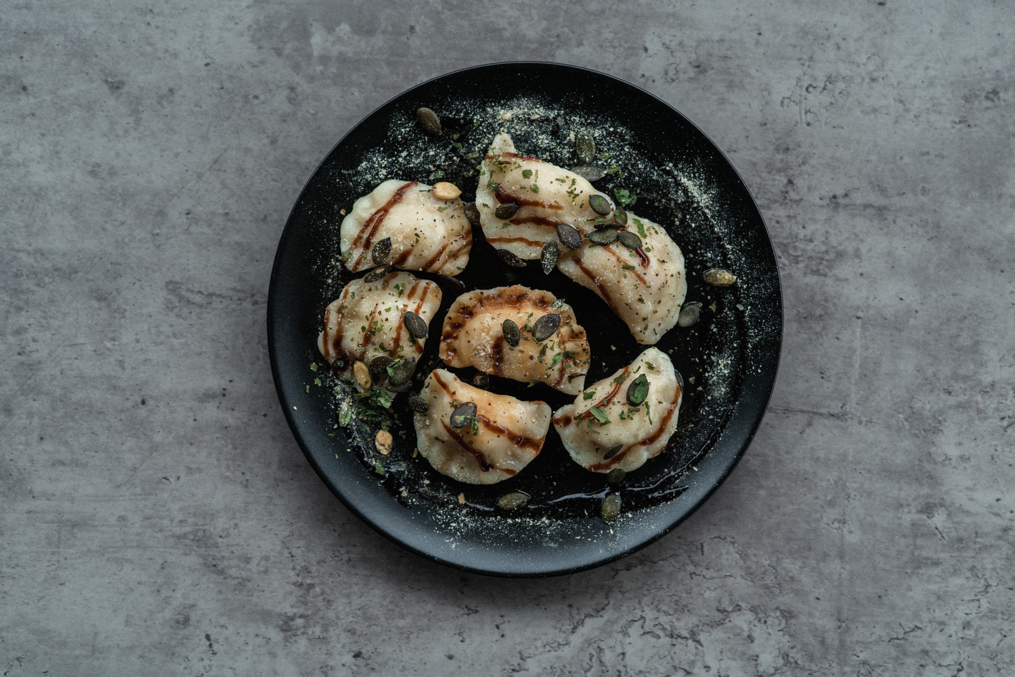 Czarny talerz z pierogami polanymi sosem, posypanymi ziołami i pestkami dyni na szarym tle.