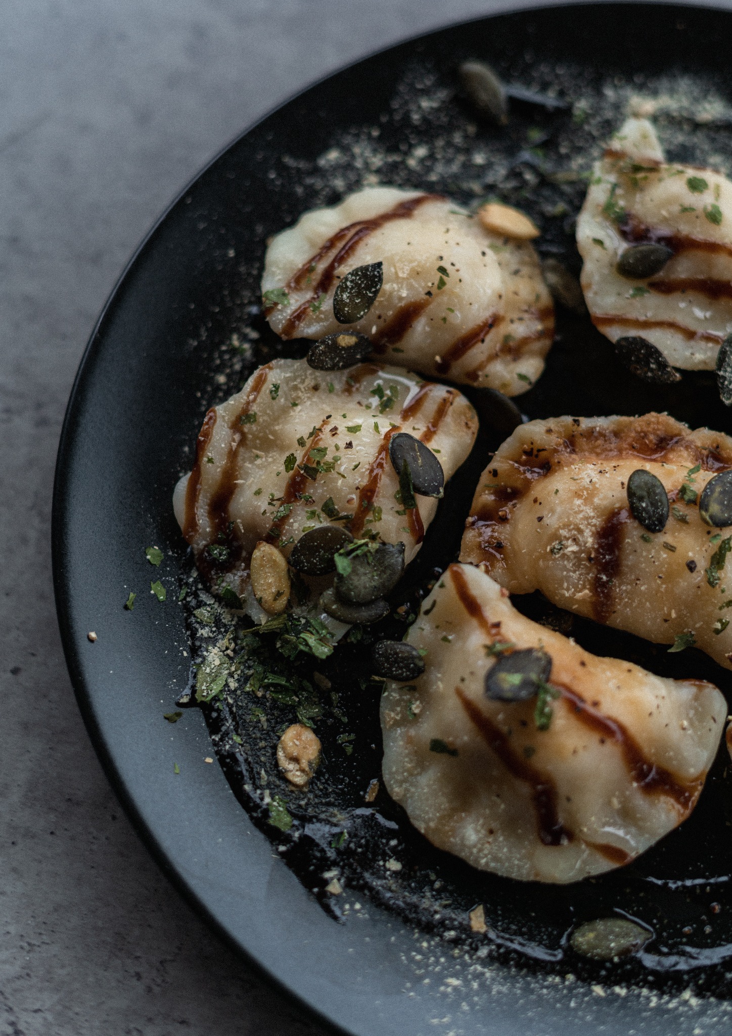 Smażone pierogi okraszone sosem balsamicznym, pestkami dyni i posypane ziołami na czarnym talerzu, widoczne z bliska na szarym tle.