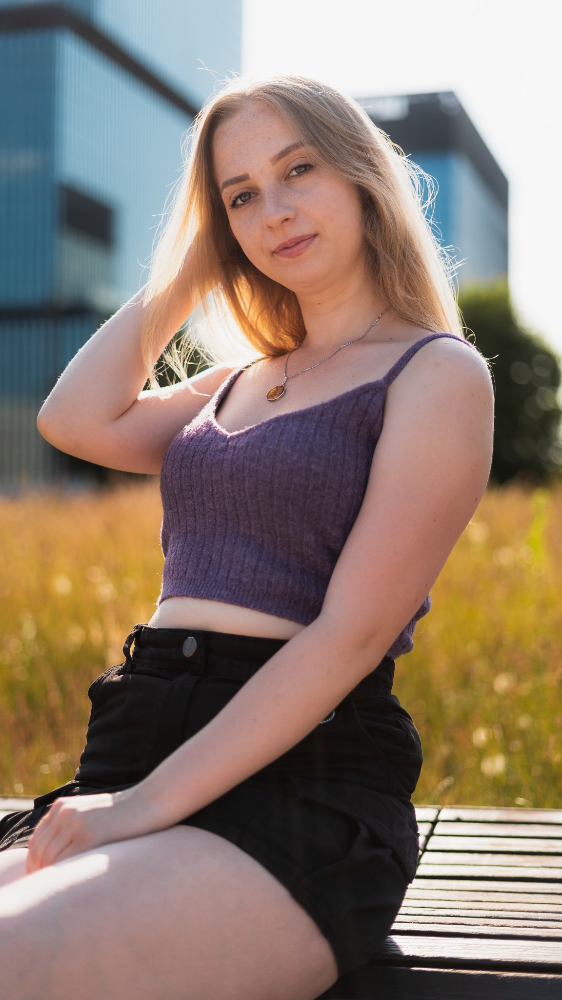 Portret młodej kobiety z piegami, siedzącej na drewnianej ławce w parku, w tle nowoczesny budynek biurowy i wysoka trawa.