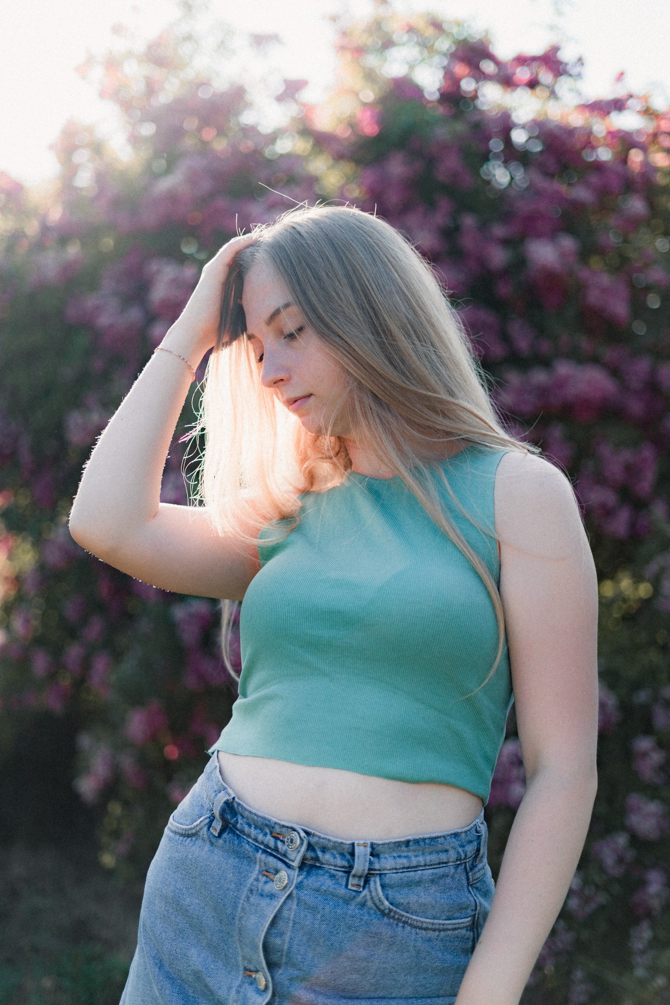 Młoda kobieta w zielonym topie i dżinsowych szortach, pozująca na tle kwitnącego drzewa w promieniach zachodzącego słońca.