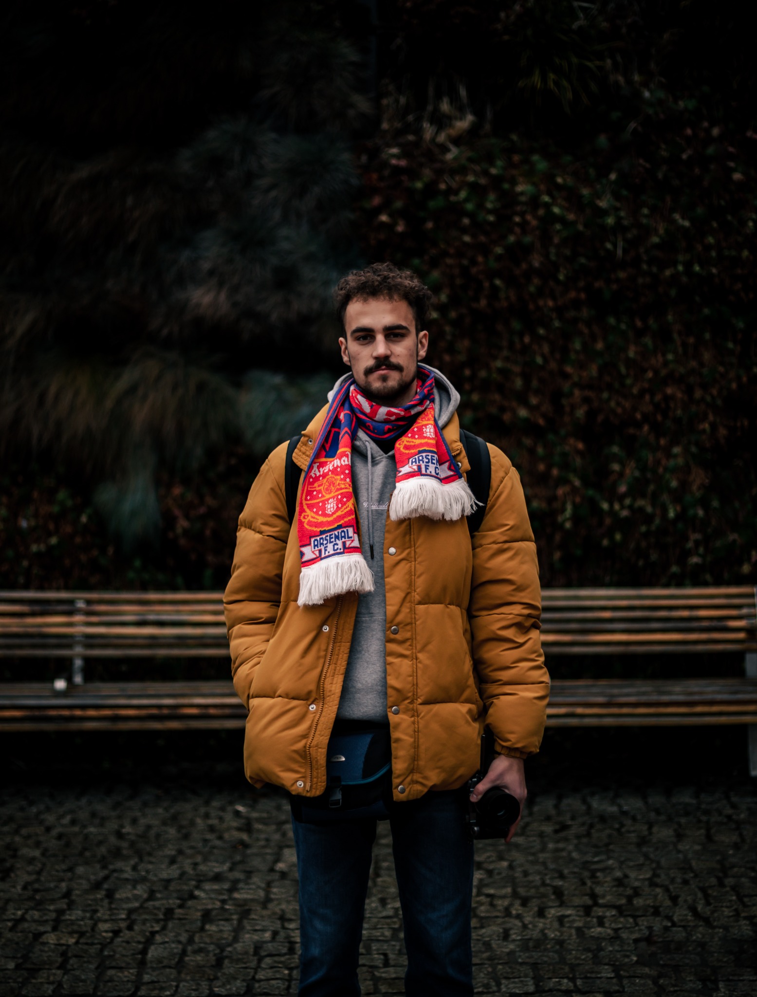 Mężczyzna z wąsami w żółtej kurtce, szarym hoodie i szaliku Arsenalu, stojący na brukowanej ścieżce przed ławką i krzewami, trzymający aparat w dłoni.