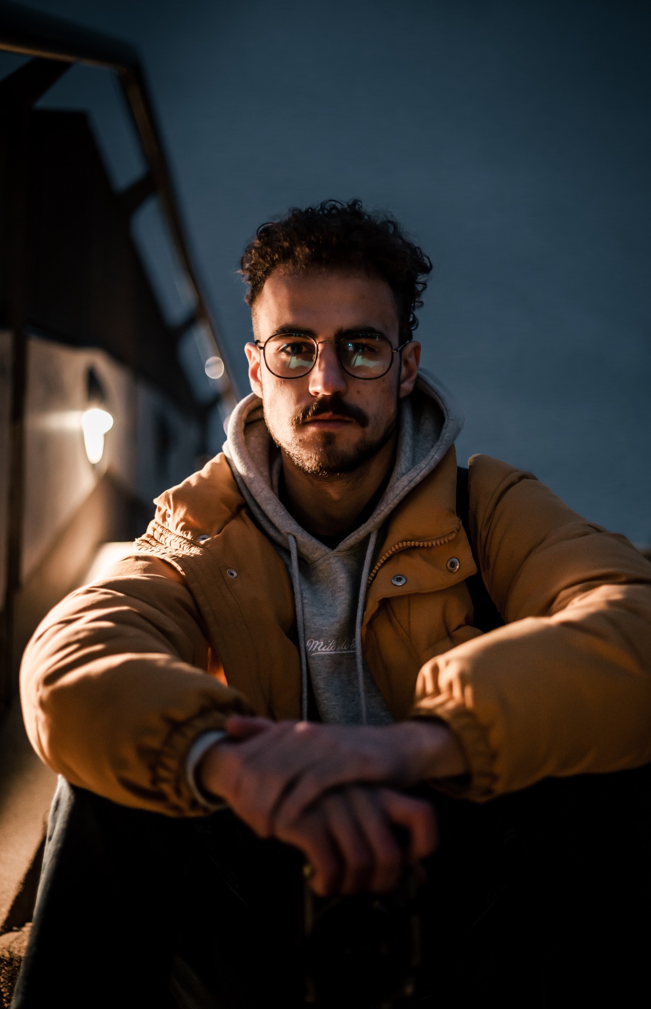 Portret młodego mężczyzny w okularach, z kręconymi włosami i brodą, ubranego w żółtą kurtkę i szarą bluzę z kapturem, siedzącego na schodach w miejskim otoczeniu o zmierzchu.