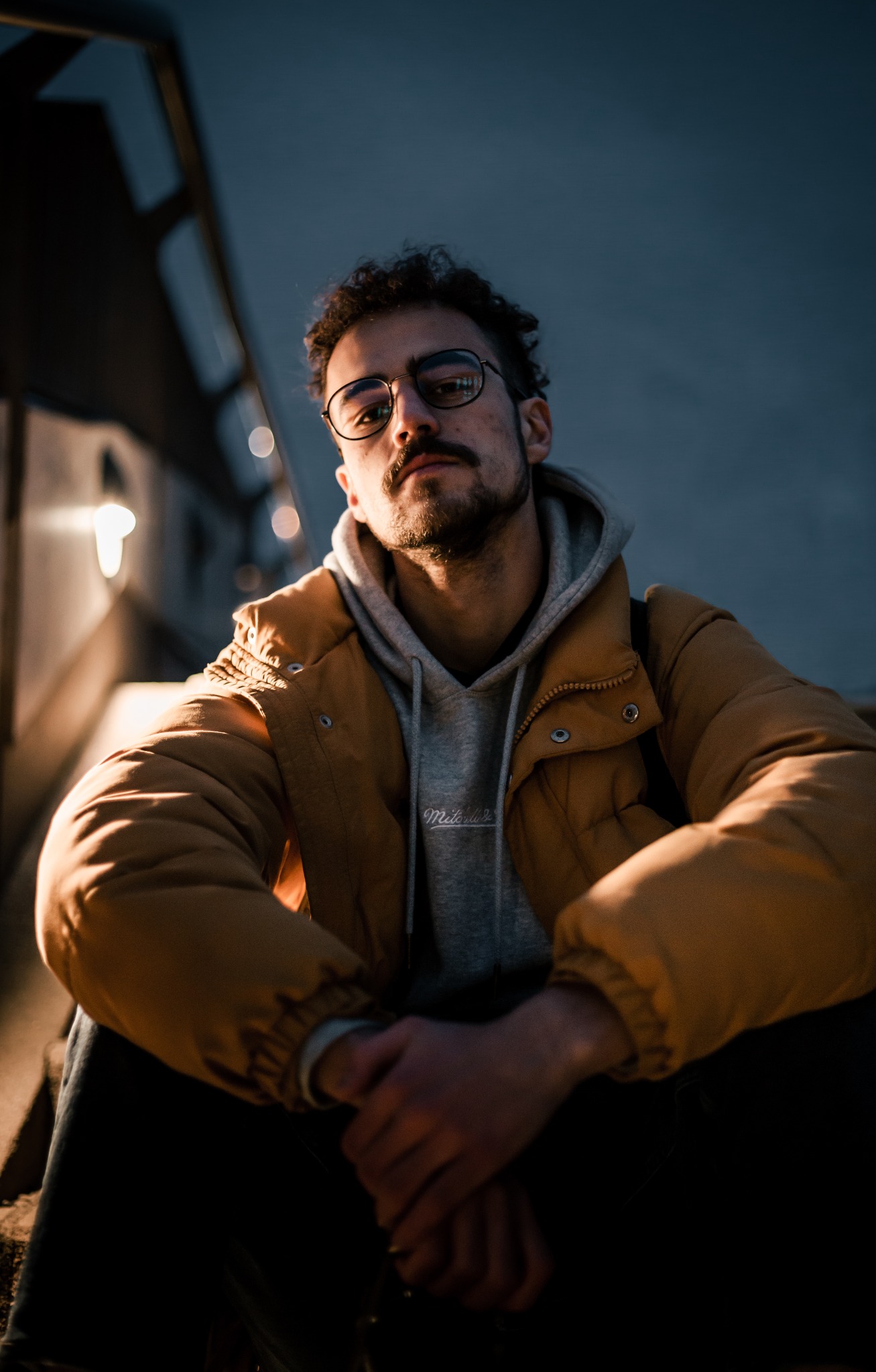 Portret młodego mężczyzny w okularach, w żółtej kurtce i szarej bluzie z kapturem, siedzącego na schodach w miejskiej scenerii, oświetlonego światłem lamp ulicznych.