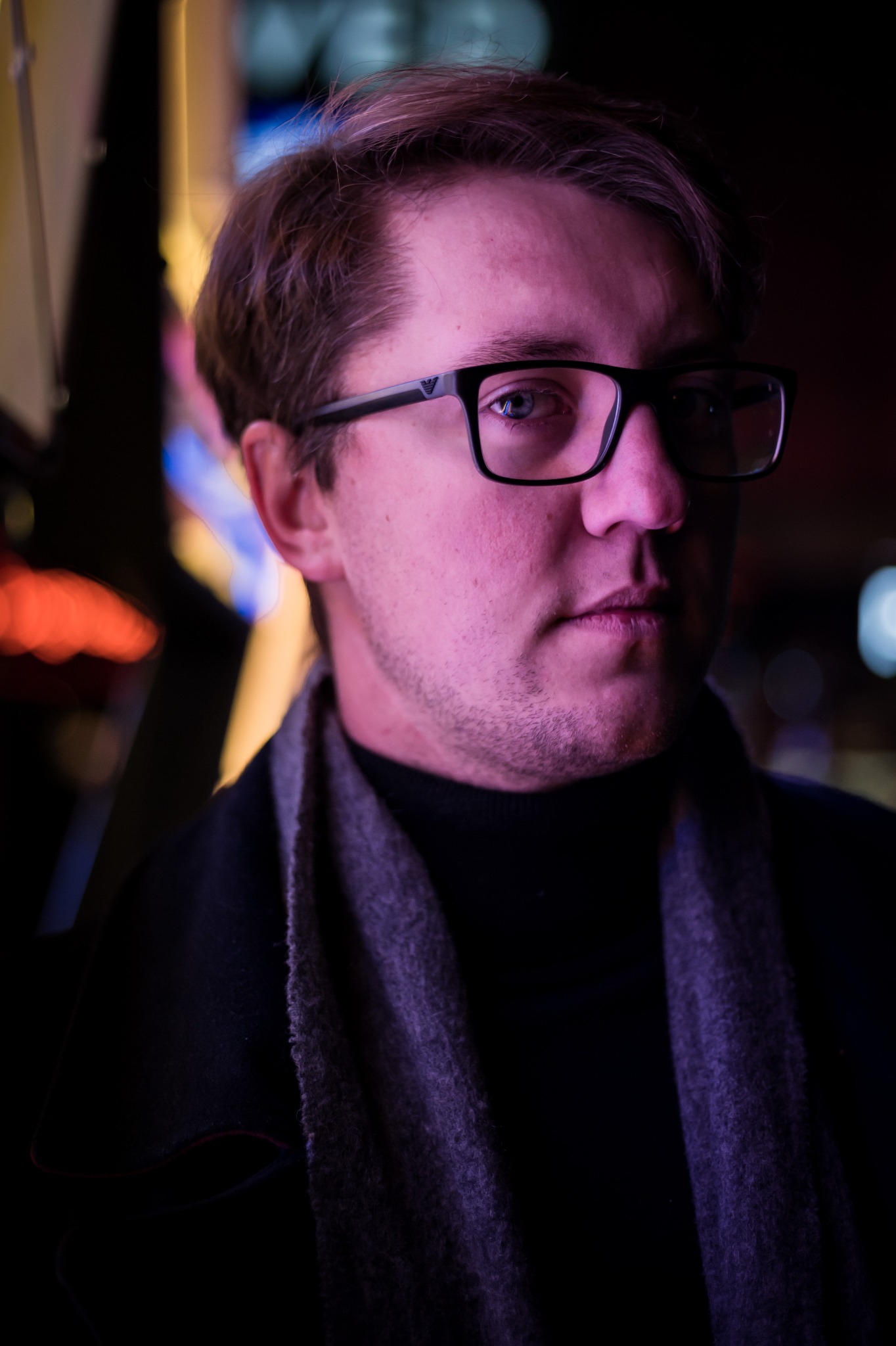 Mężczyzna w okularach i szaliku, oświetlony neonowym, fioletowym światłem, z widocznym fragmentem neonowego szyldu w tle.