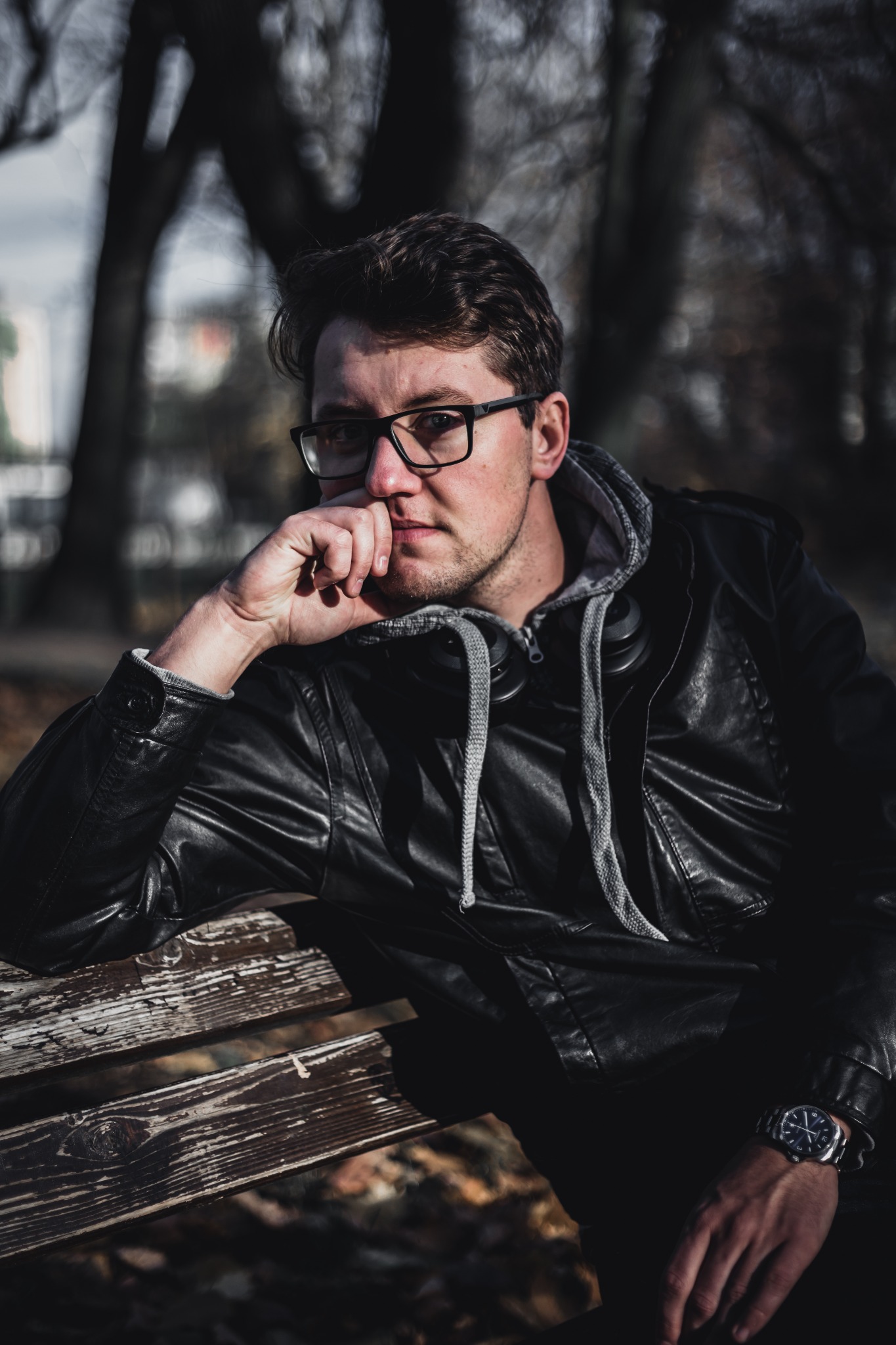 Młody mężczyzna w okularach, czarnej skórzanej kurtce i szarej bluzie z kapturem, siedzi na drewnianej ławce w parku, zamyślony, z ręką podpartą na brodzie, widoczny zegarek na nadgarstku.