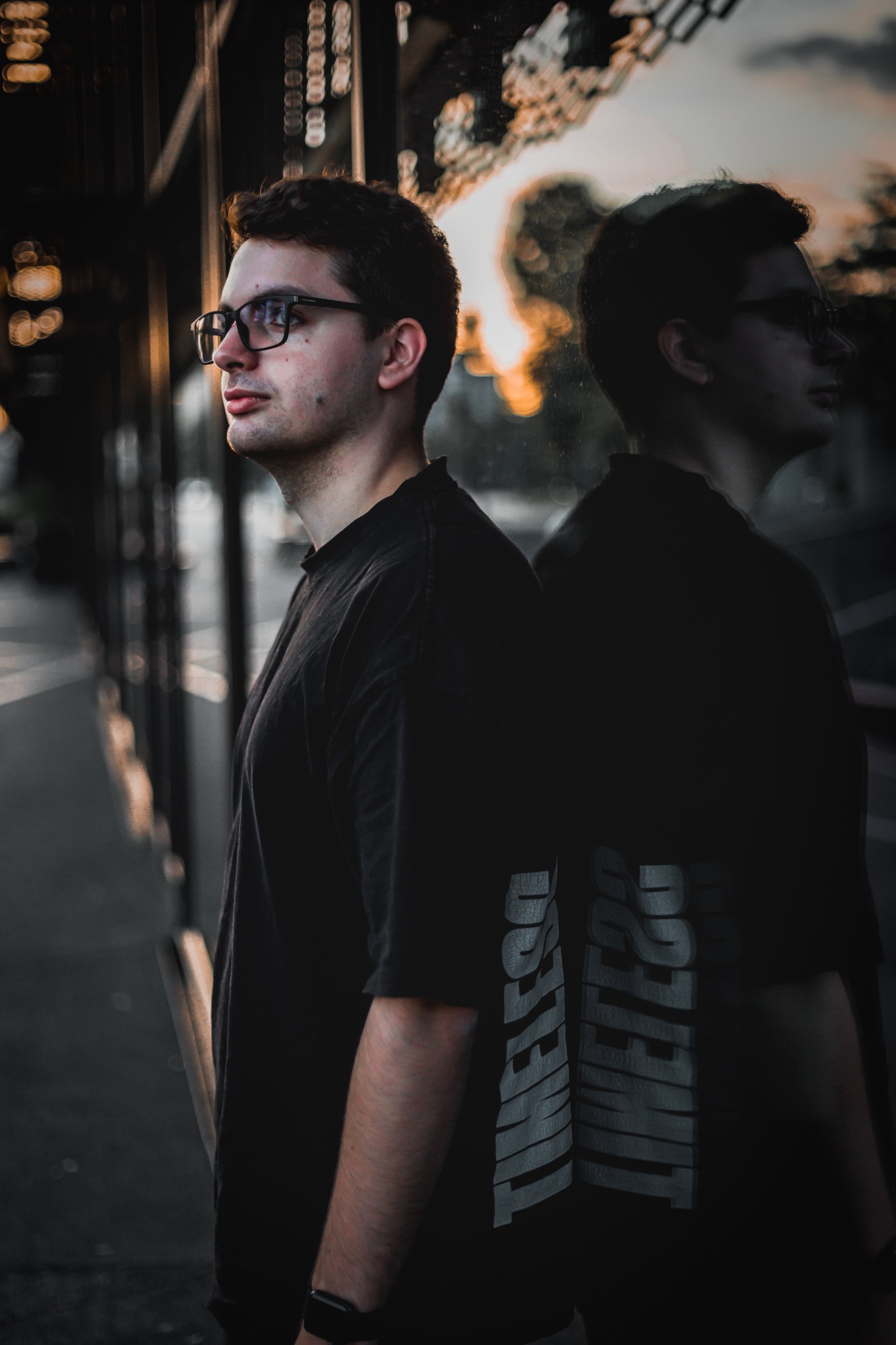 Portret młodego mężczyzny w okularach, opierającego się o szklaną powierzchnię z odbiciem, ubrany w czarny t-shirt z napisem 'TIMELESS', na tle miejskiego krajobrazu z elementami zachodzącego słońca.