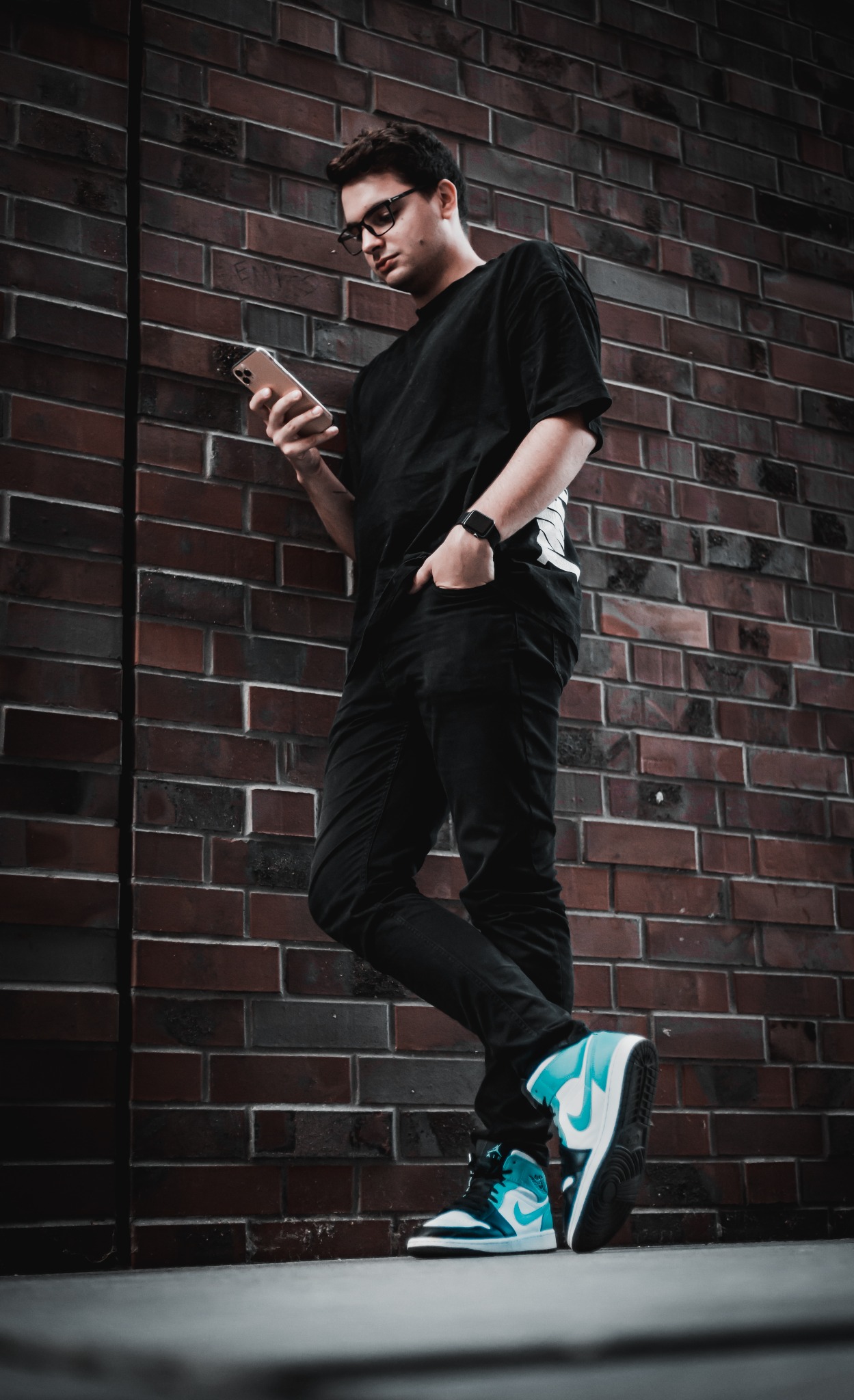 Młody mężczyzna w okularach, ubrany na czarno, opiera się o ceglaną ścianę, przeglądając coś na smartfonie. Na nogach ma biało-turkusowe sneakersy.