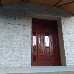 Wejście do domu z elewacją wykończoną szarym kamieniem, ciemnoczerwone drzwi z przeszkleniami, skrzynka elektryczna na ścianie.