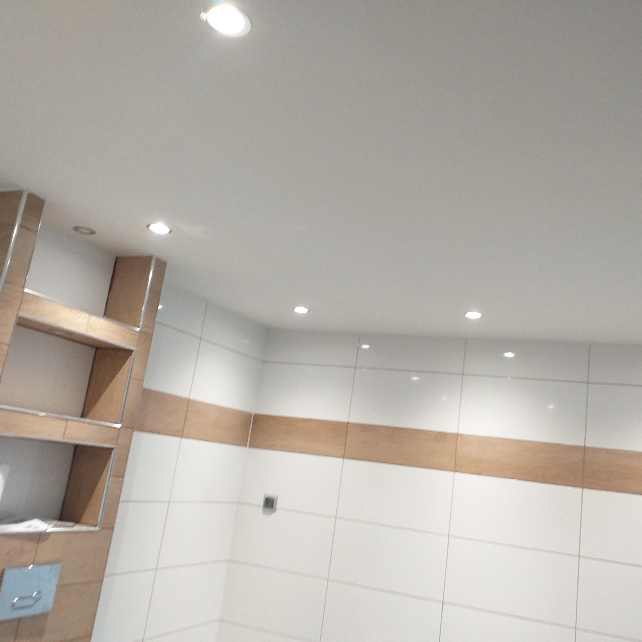 Wykończona łazienka z białymi płytkami i drewnianymi akcentami, widoczne oświetlenie sufitowe i półki wnękowe z drewnianym obramowaniem.