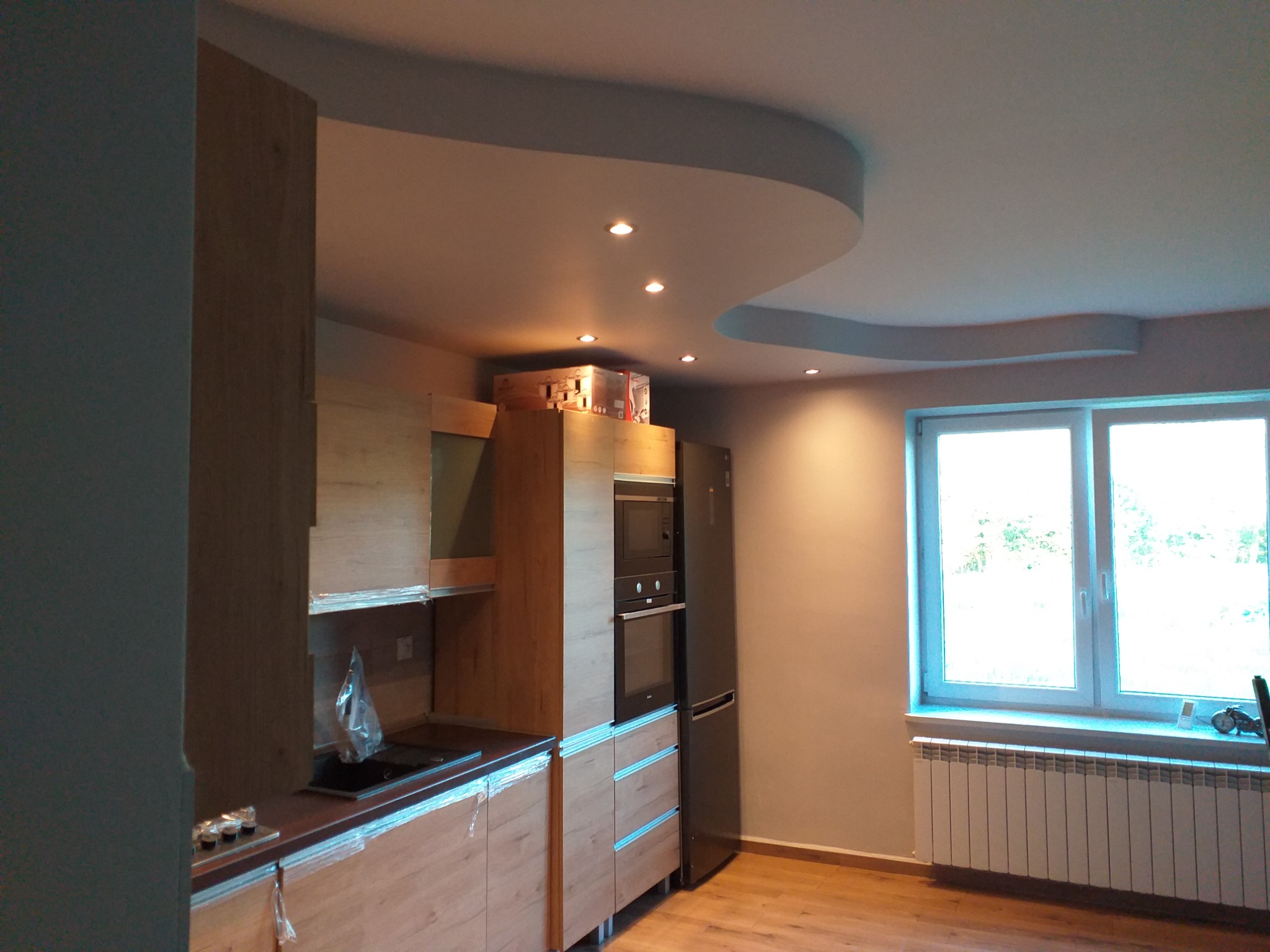 Wnętrze kuchni w trakcie prac wykończeniowych; widoczne meble kuchenne z okleiną drewnopodobną, sprzęt AGD w zabudowie, podwieszany sufit z oświetleniem punktowym oraz okno z grzejnikiem.