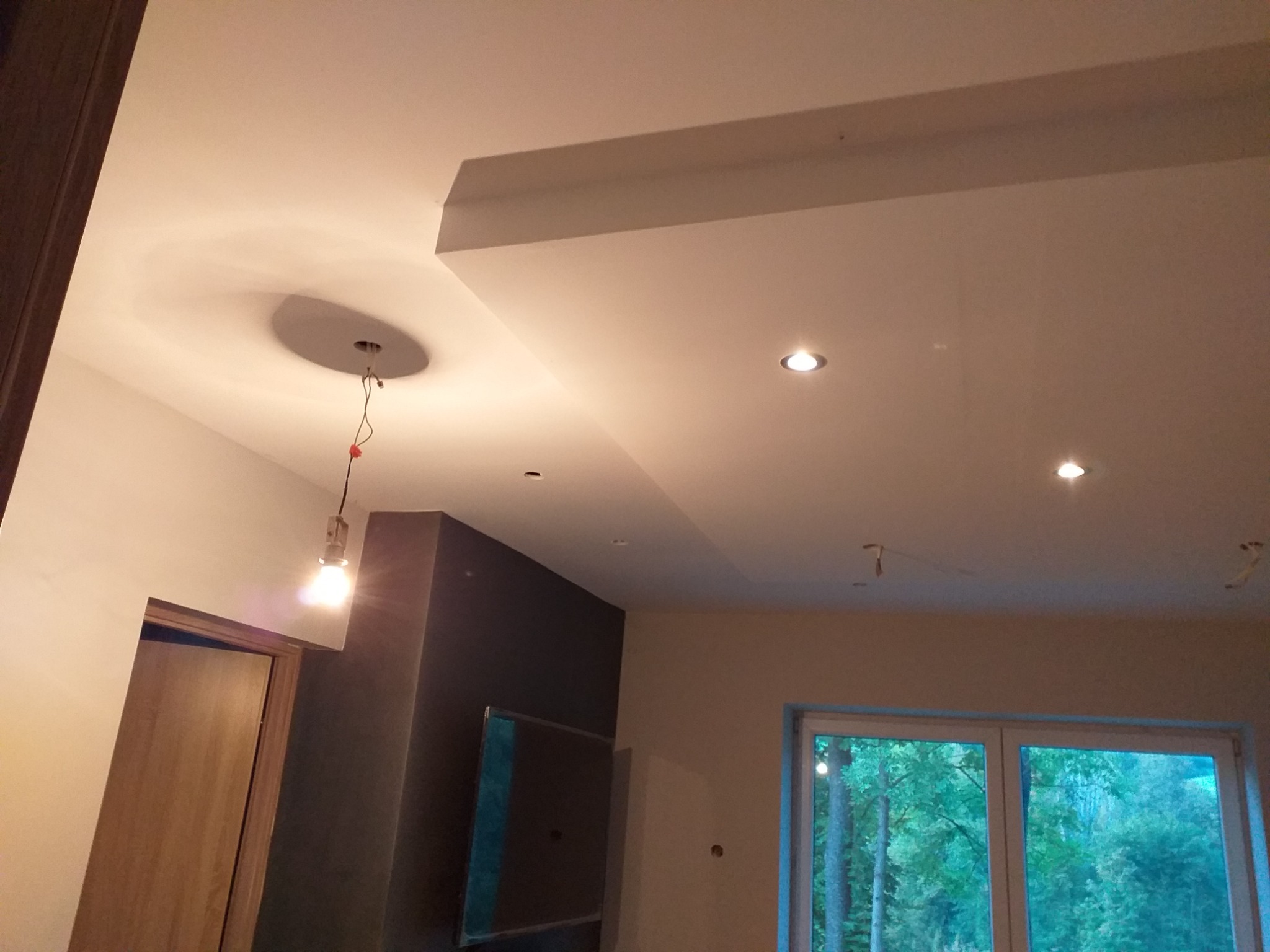 Fragment wnętrza w trakcie prac wykończeniowych, widoczny sufit z podwieszanymi elementami i punktowym oświetleniem, wisząca żarówka na kablu oraz okno z widokiem na zieleń.