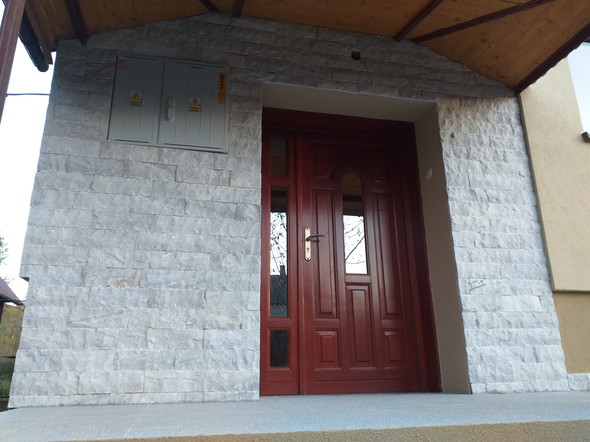 Wejście do domu z elewacją wykończoną szarym kamieniem, ciemnoczerwone drzwi z przeszkleniami, skrzynka elektryczna na ścianie.