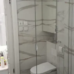 Nowoczesna łazienka z marmurowymi płytkami na ścianach i czarną, marmurkową podłogą, wisząca toaleta z ukrytą spłuczką oraz szklana kabina prysznicowa bez brodzika.