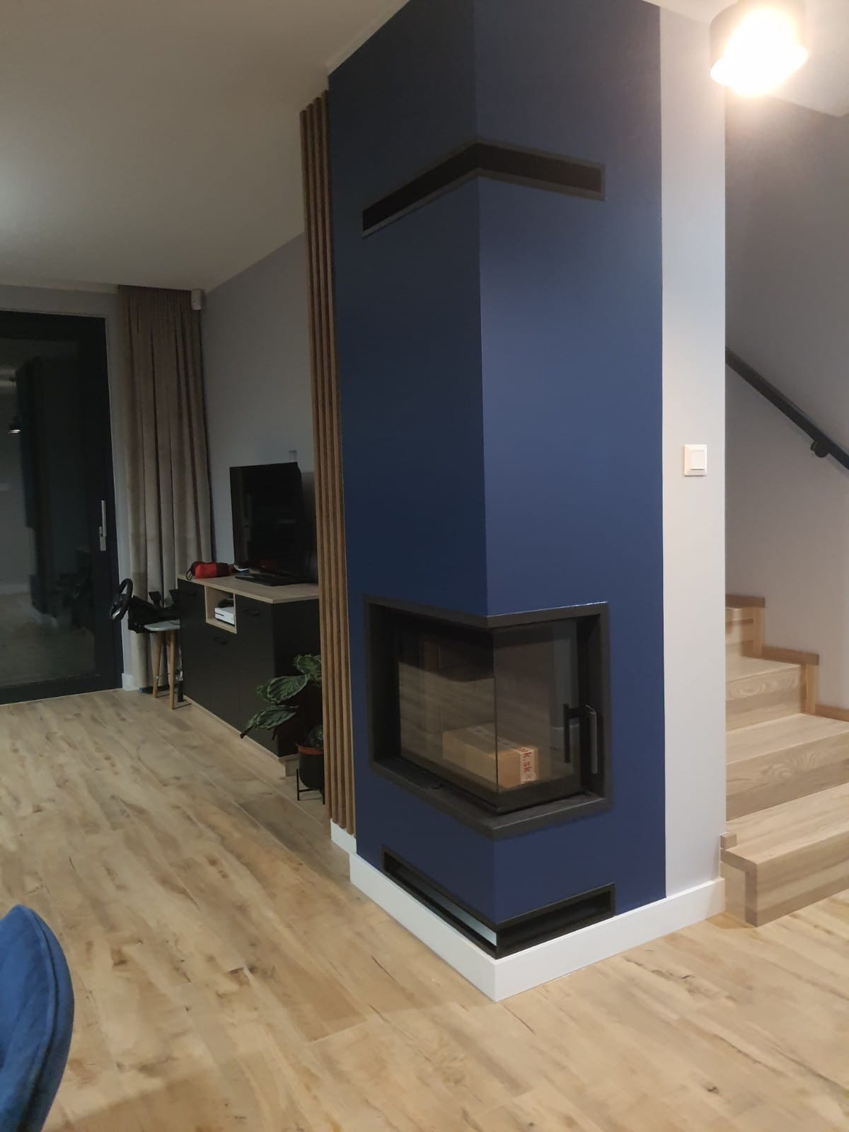 Nowoczesny salon z drewnianą podłogą, niebieską ścianą z wbudowanym kominkiem i drewnianymi panelami pionowymi. W tle widoczny telewizor na szafce i schody z drewnianymi stopniami.