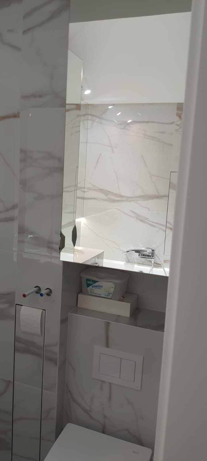 Nowoczesna łazienka w trakcie wykańczania, widoczne marmurowe płytki, zabudowana toaleta z przyciskiem spłukującym, wystające rury instalacji wodnej oraz półka z akcesoriami.