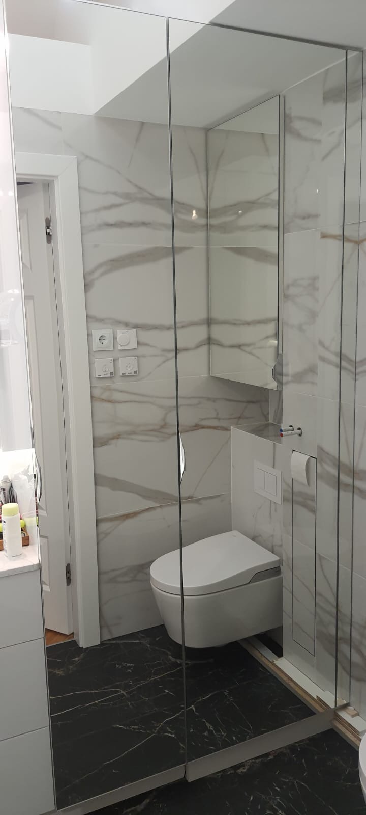 Nowoczesna łazienka z marmurowymi płytkami na ścianach i czarną, marmurkową podłogą, wisząca toaleta z ukrytą spłuczką oraz szklana kabina prysznicowa bez brodzika.