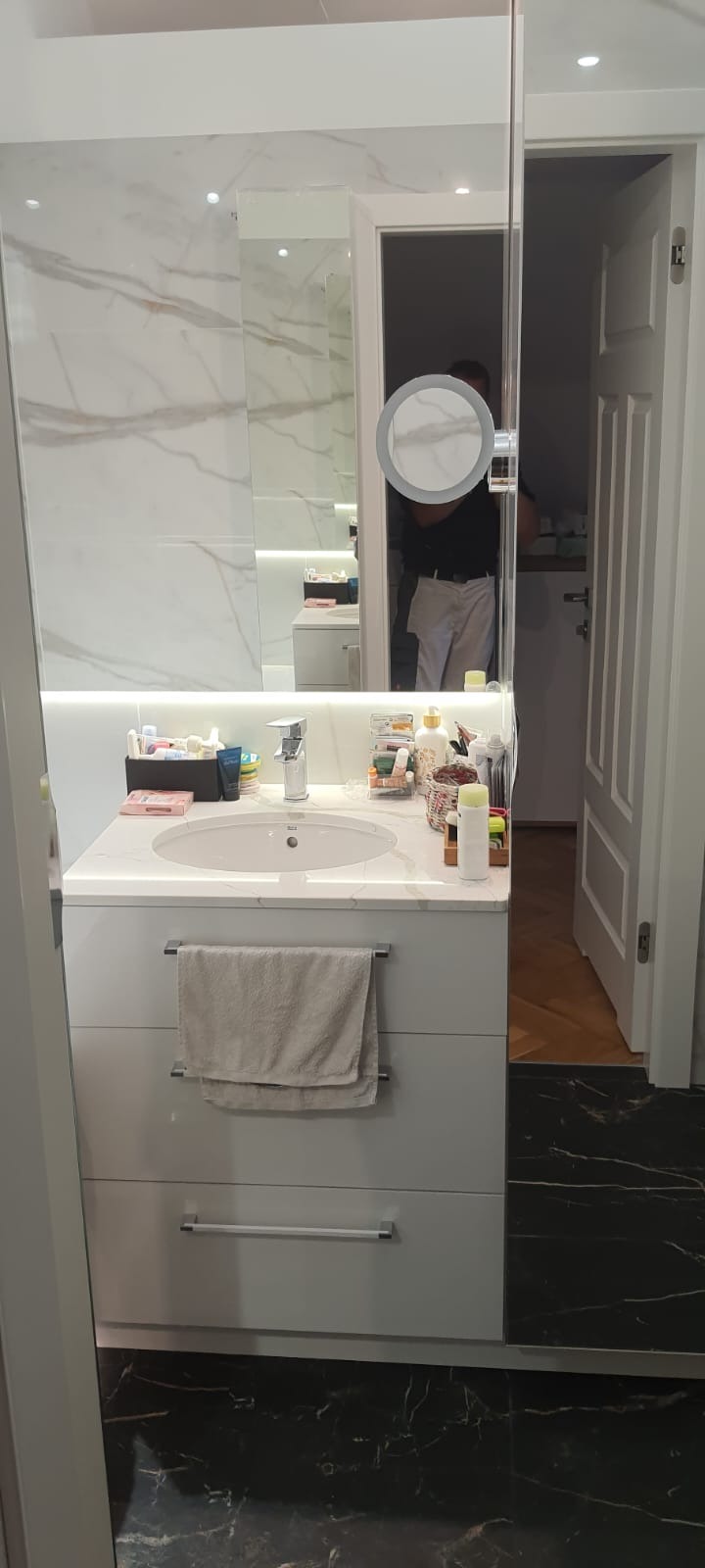 Nowoczesna łazienka z marmurową ścianą, podświetlanym lustrem i umywalką nablatową na szafce z szufladami, z widocznym fragmentem otwartych drzwi do innego pomieszczenia z parkietem.