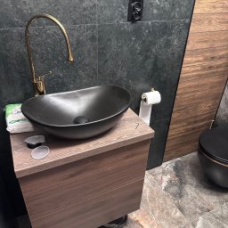 JEL-TOMIX TOMASZ CIURA - Nowoczesna łazienka z czarną umywalką na drewnianej szafce, złotą baterią i marmurową podłogą. Na ścianie ciemne płytki i drewniane panele.