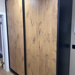 JEL-TOMIX TOMASZ CIURA - Szafa przesuwna z drewnopodobnymi drzwiami i czarnymi obramowaniami, widoczna z boku, z fragmentem podłogi i drzwi w tle.