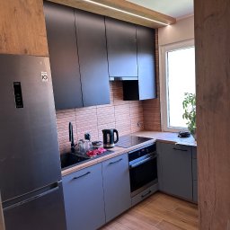 JEL-TOMIX TOMASZ CIURA - Nowoczesna kuchnia z szarymi szafkami, czarnymi frontami górnymi i drewnianymi akcentami. Widoczny sprzęt AGD: lodówka LG, płyta indukcyjna i piekarnik. Oświetlenie LED pod szafkami.