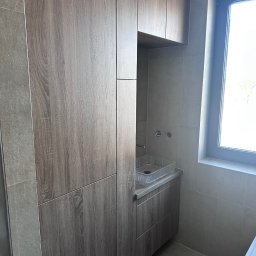 JEL-TOMIX TOMASZ CIURA - Nowoczesna łazienka z zabudową meblową w odcieniach szarości i drewna. Umywalka z przezroczystego tworzywa, okno z widokiem na zewnątrz. Minimalistyczny design.