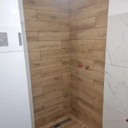 Wykończony fragment kabiny prysznicowej z drewnopodobnymi płytkami na ścianach i podłodze, widoczne przyłącza wodne i odpływ liniowy.