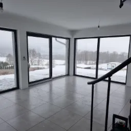 Wnętrze jasnego, minimalistycznego pokoju z dużymi oknami panoramicznymi, przez które widać zimowy krajobraz. Na pierwszym planie fragment czarnej, spiralnej klatki schodowej. Na podłodze szara...