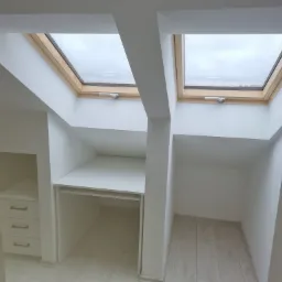 Białe wnętrze poddasza z dwoma oknami dachowymi, zabudową meblową obejmującą biurko i komodę z trzema szufladami, widoczne drewniane wykończenie okien i panele podłogowe.