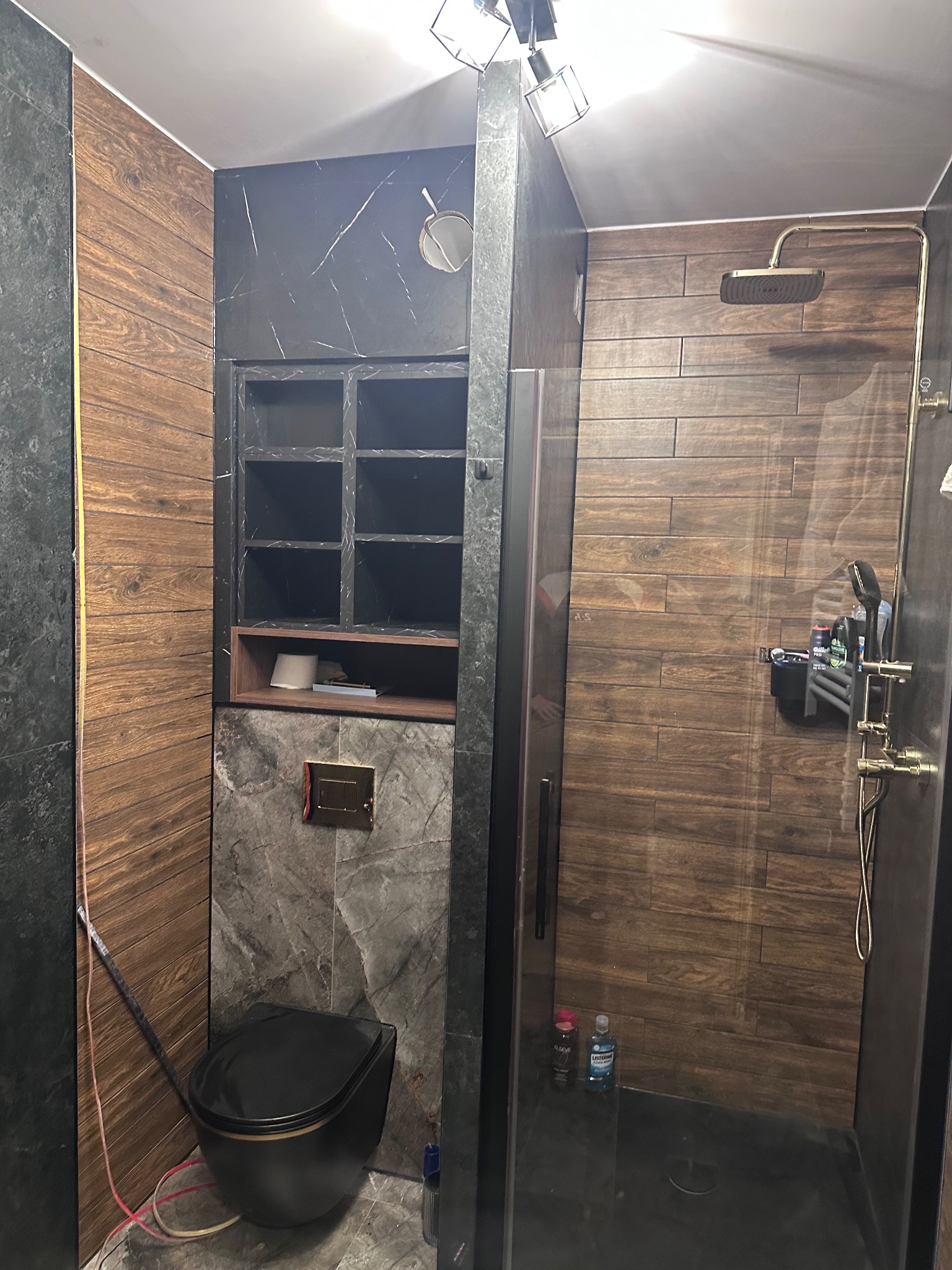 Nowoczesna łazienka z kabiną prysznicową typu walk-in, drewnopodobnymi płytkami i czarną armaturą. Widoczna toaleta w kolorze czarnym i półki wnękowe.