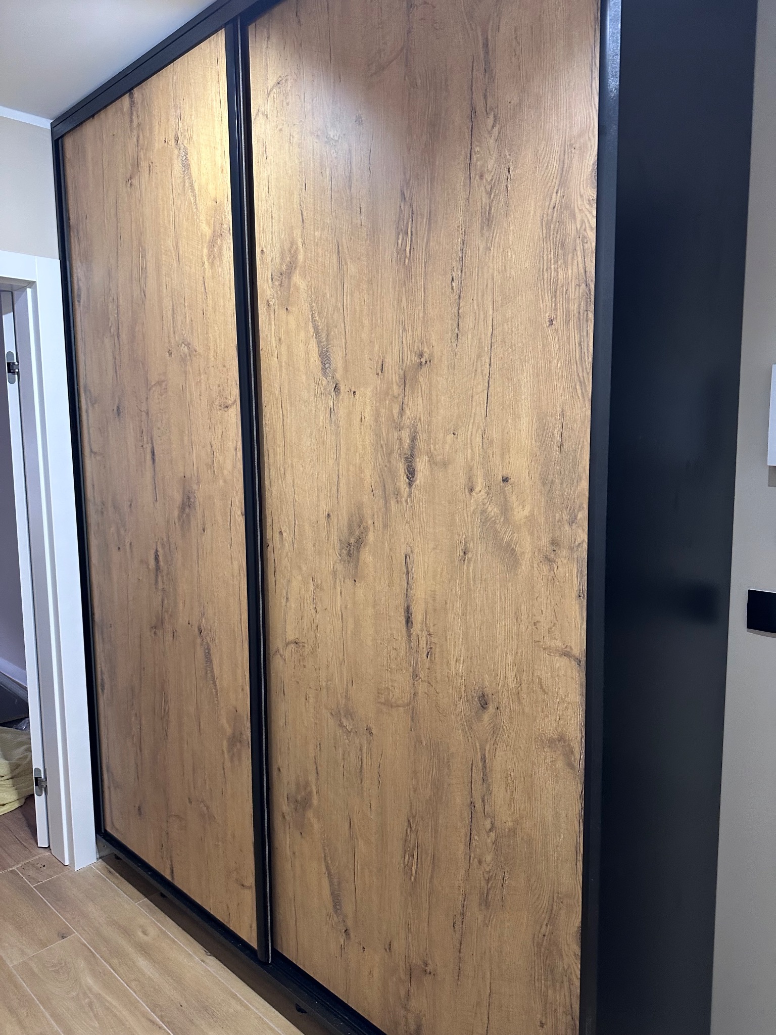 Szafa przesuwna z drewnopodobnymi drzwiami i czarnymi obramowaniami, widoczna z boku, z fragmentem podłogi i drzwi w tle.