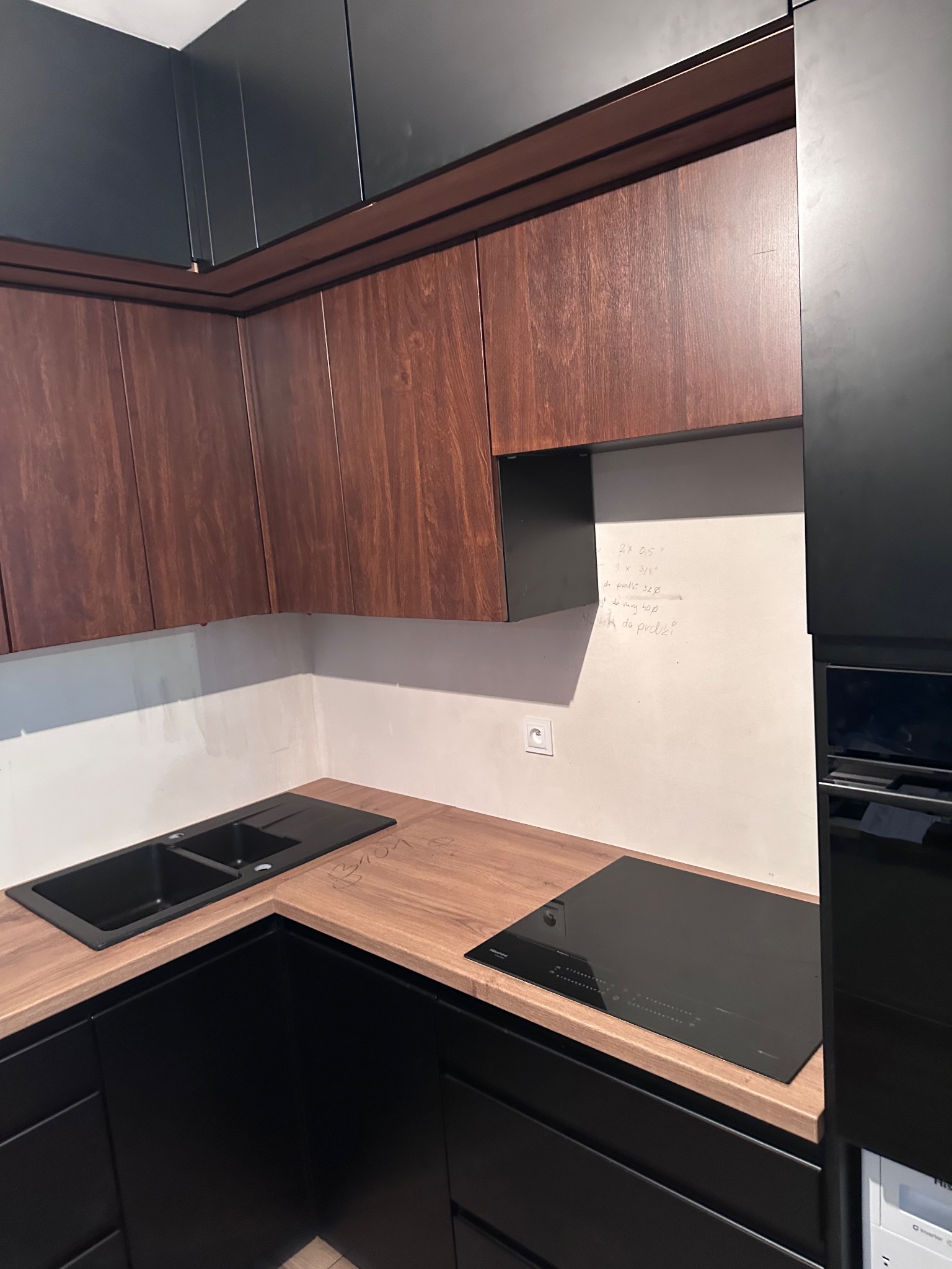 Nowoczesna kuchnia z drewnianymi szafkami górnymi i czarnymi dolnymi. Na blacie czarny zlew i płyta indukcyjna. Na ścianie notatki z wymiarami.