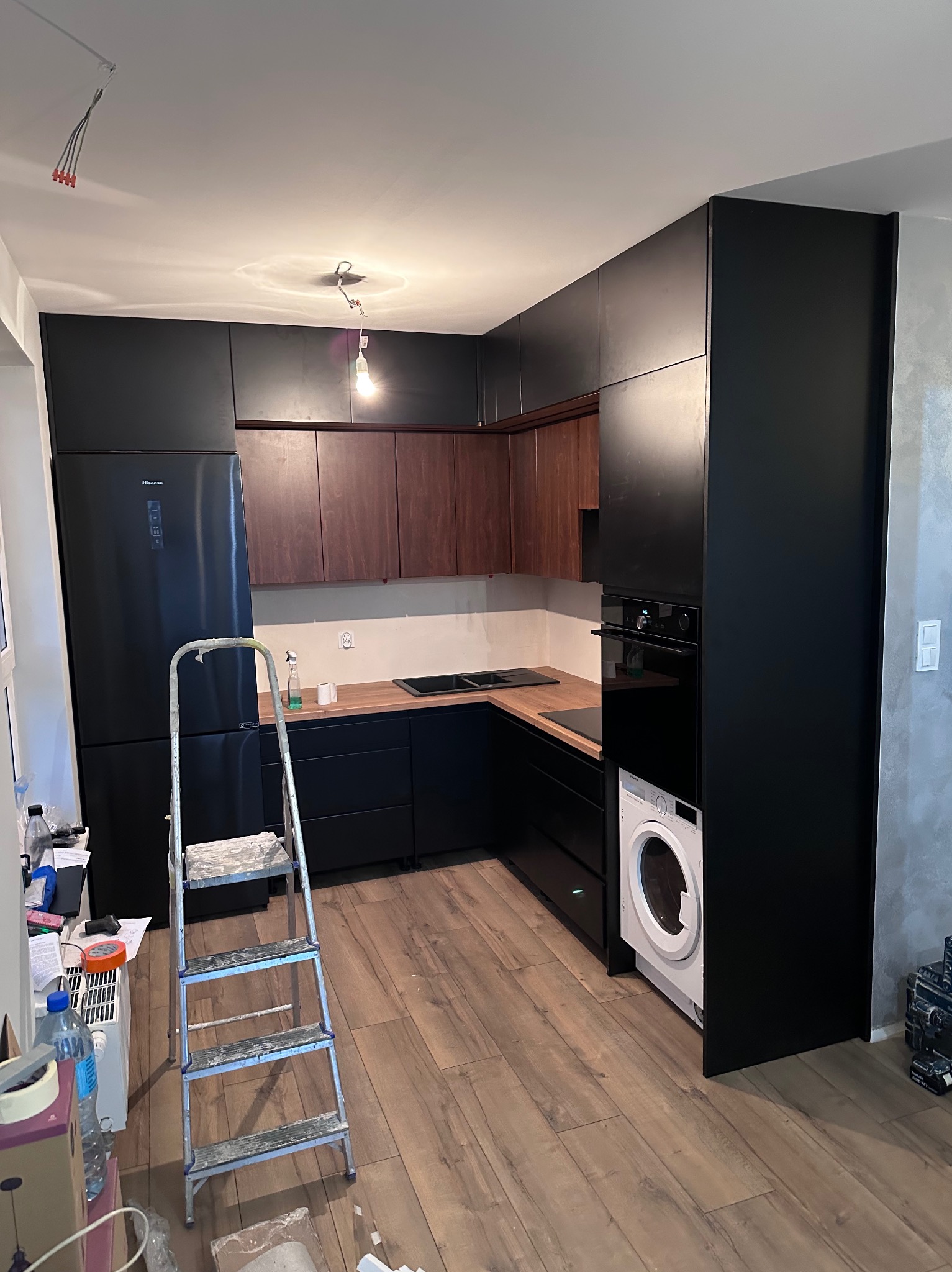 Częściowo wykończona kuchnia z czarnymi i drewnianymi szafkami, sprzętem AGD i drabiną. Widoczne niedokończone elementy instalacji elektrycznej.