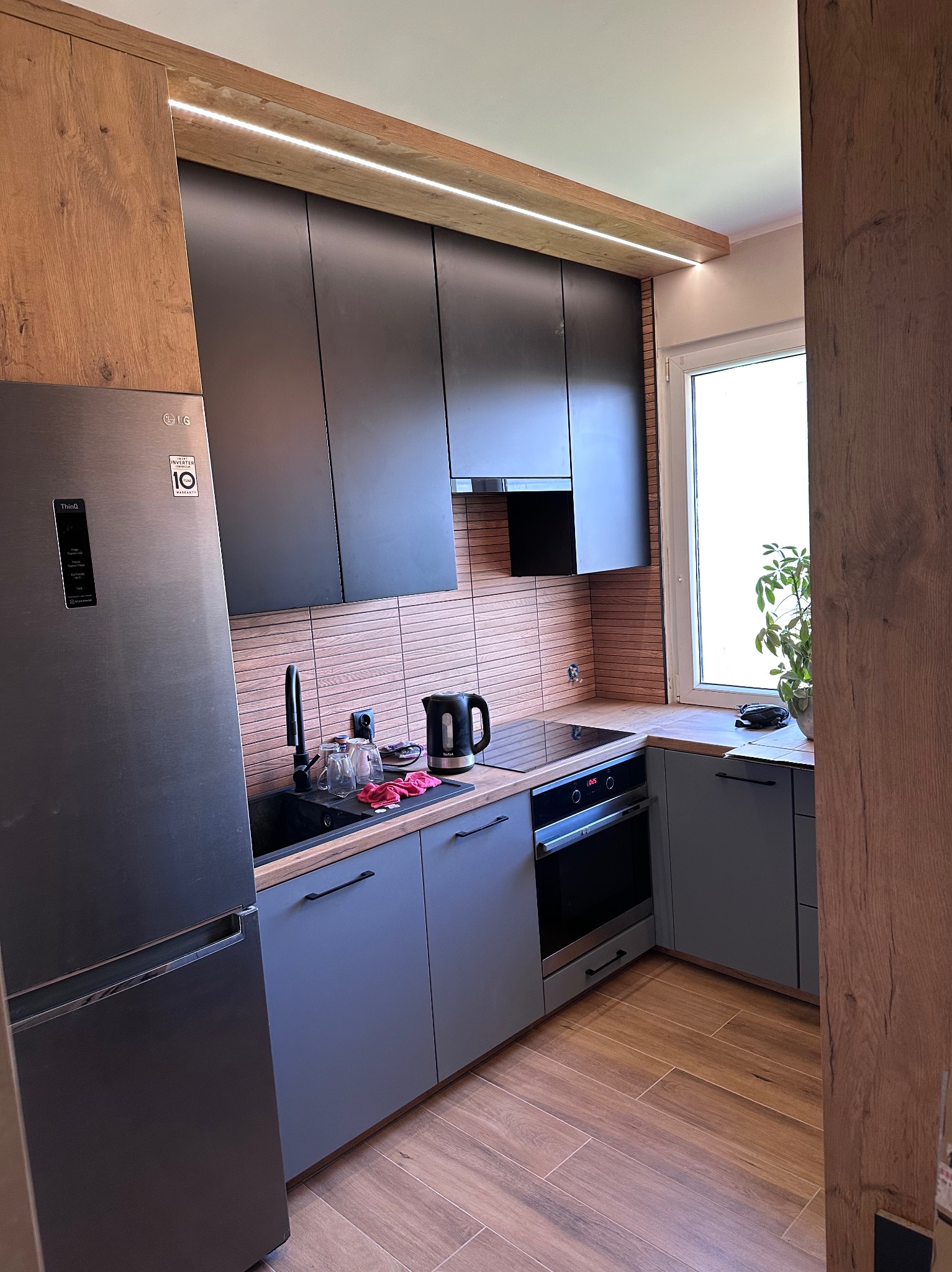 Nowoczesna kuchnia z szarymi szafkami, czarnymi frontami górnymi i drewnianymi akcentami. Widoczny sprzęt AGD: lodówka LG, płyta indukcyjna i piekarnik. Oświetlenie LED pod szafkami.