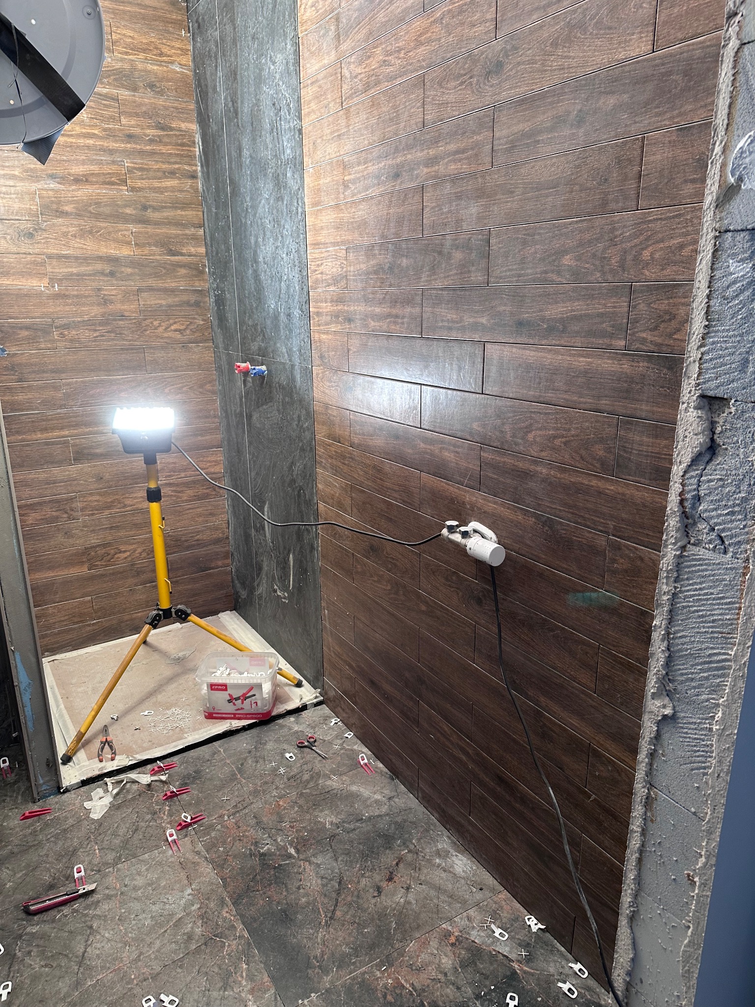 Układanie płytek imitujących drewno na ścianie w łazience, widoczne narzędzia i system poziomowania płytek. Oświetlenie halogenowe na statywie. Szare płytki na podłodze.