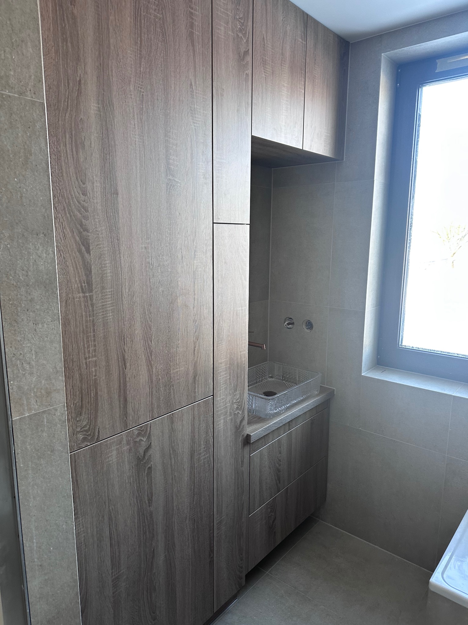 Nowoczesna łazienka z zabudową meblową w odcieniach szarości i drewna. Umywalka z przezroczystego tworzywa, okno z widokiem na zewnątrz. Minimalistyczny design.