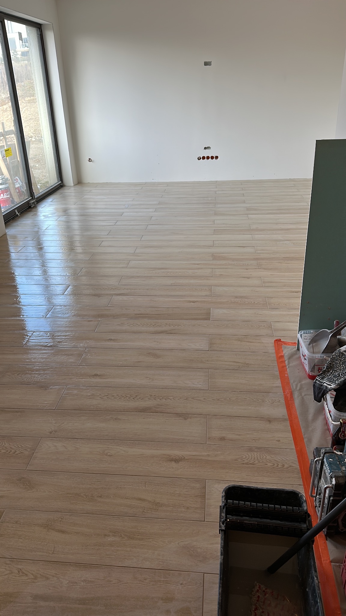 Ułożona podłoga z płytek imitujących drewno w jasnym wnętrzu. Widoczne narzędzia i akcesoria do układania płytek, duże okno z widokiem na zewnątrz.