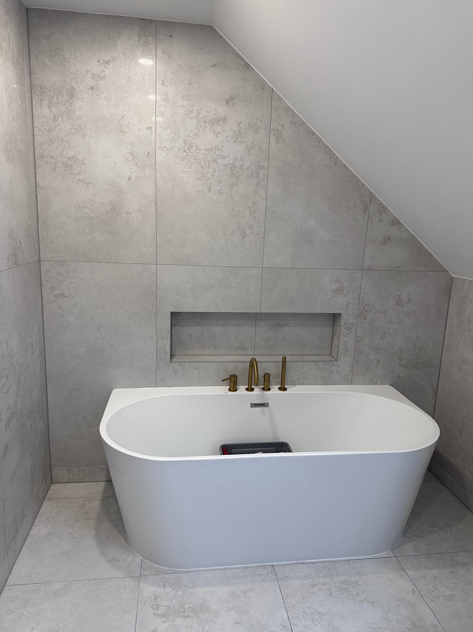 Nowoczesna łazienka z wolnostojącą wanną na tle szarych płytek imitujących beton, złota armatura i wnęka w ścianie. Minimalistyczny design.