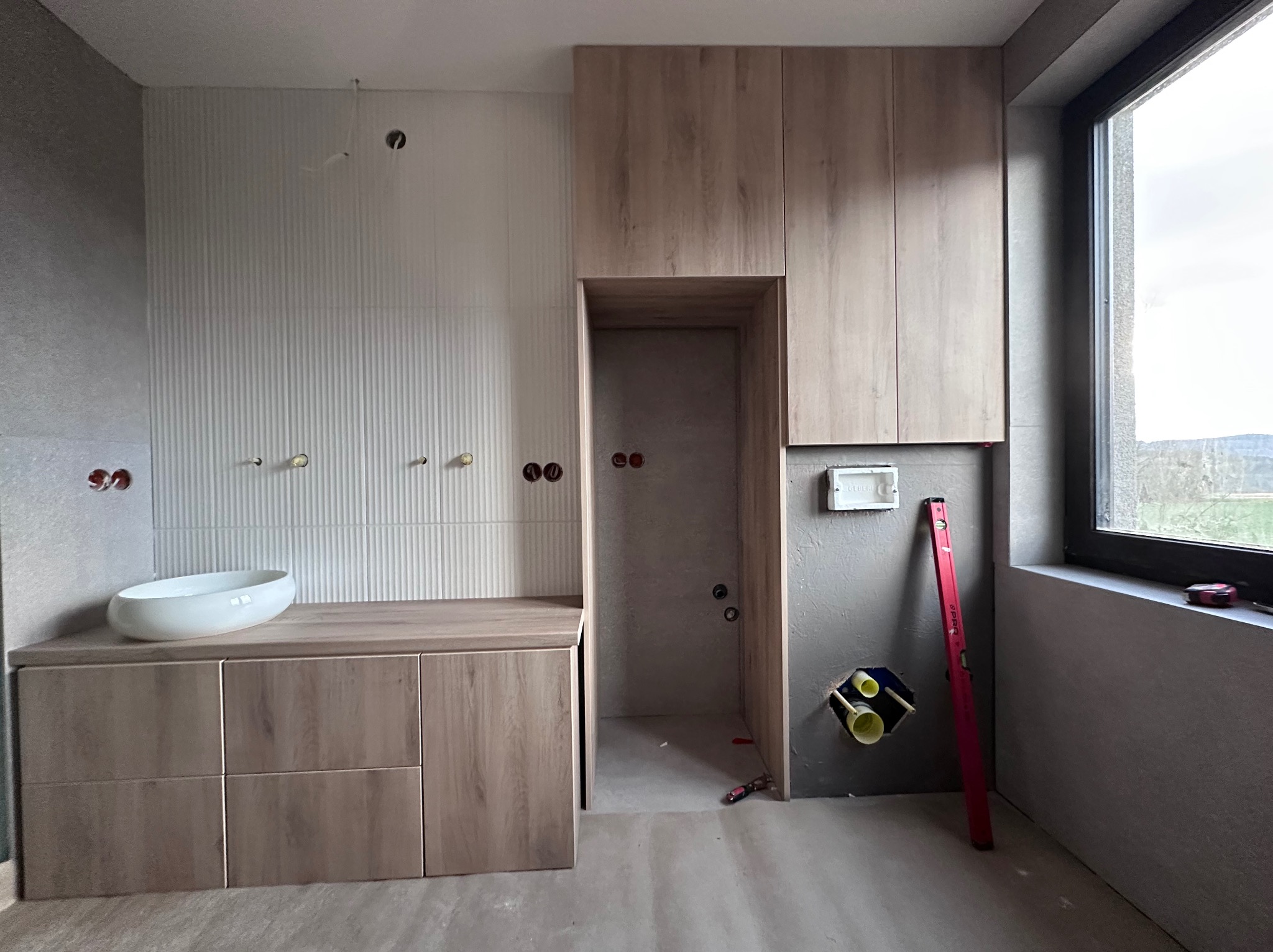 Montaż szafek łazienkowych z drewnianym fornirem, biała umywalka nablatowa, białe płytki ścienne z pionowymi żłobieniami, poziomica oparta o ścianę, surowy stan wykończenia.