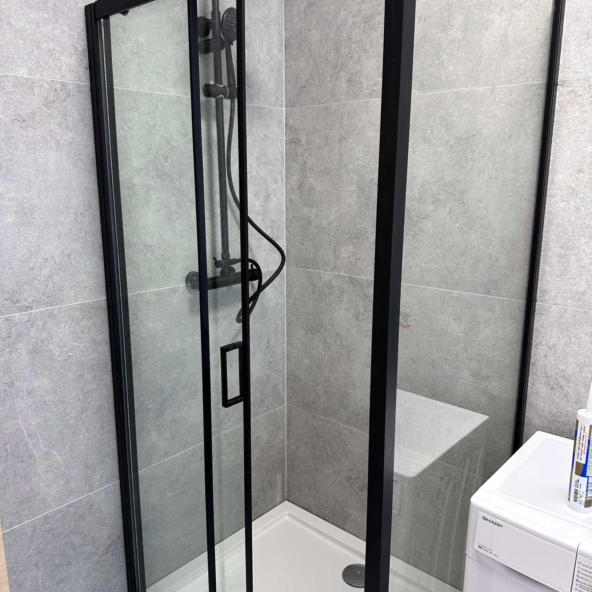Nowoczesna kabina prysznicowa z czarną ramą i deszczownicą, wykończona szarymi płytkami. Minimalistyczny design, widoczna pralka w tle.