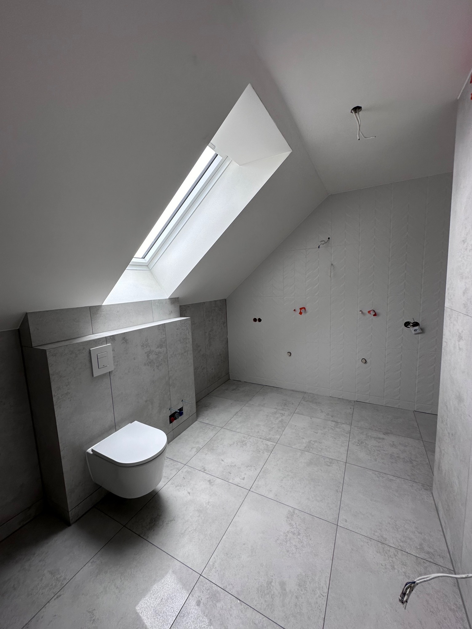 Wykończona łazienka na poddaszu w Jeżowie Sudeckim. Szare płytki, podwieszana toaleta, okno dachowe. Widoczne instalacje elektryczne i hydrauliczne.
