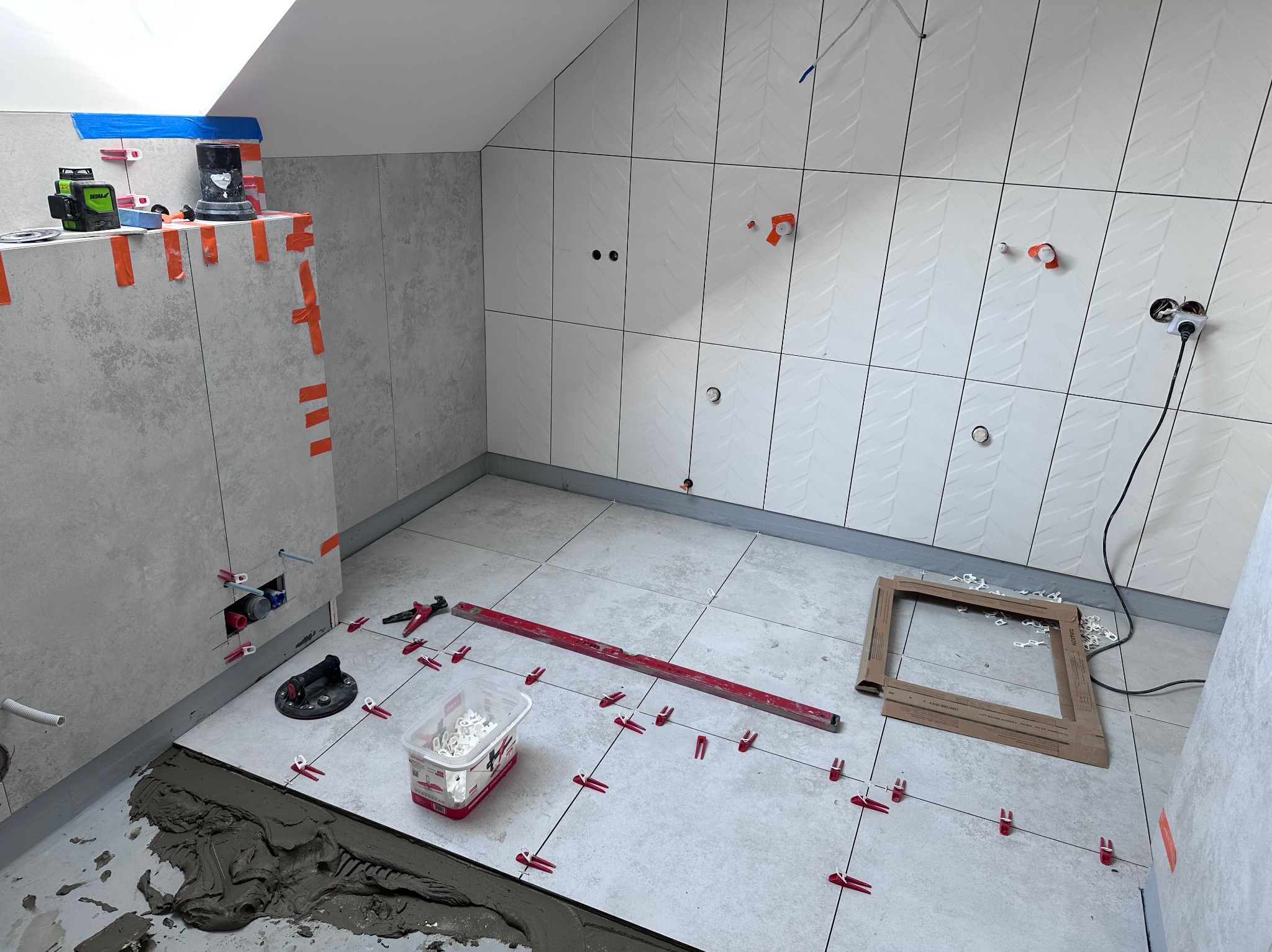 Układanie płytek w łazience na poddaszu. Widoczne narzędzia, poziomica, krzyżyki dystansowe i laser krzyżowy. Ściany wyłożone białymi płytkami z wzorem jodełki.