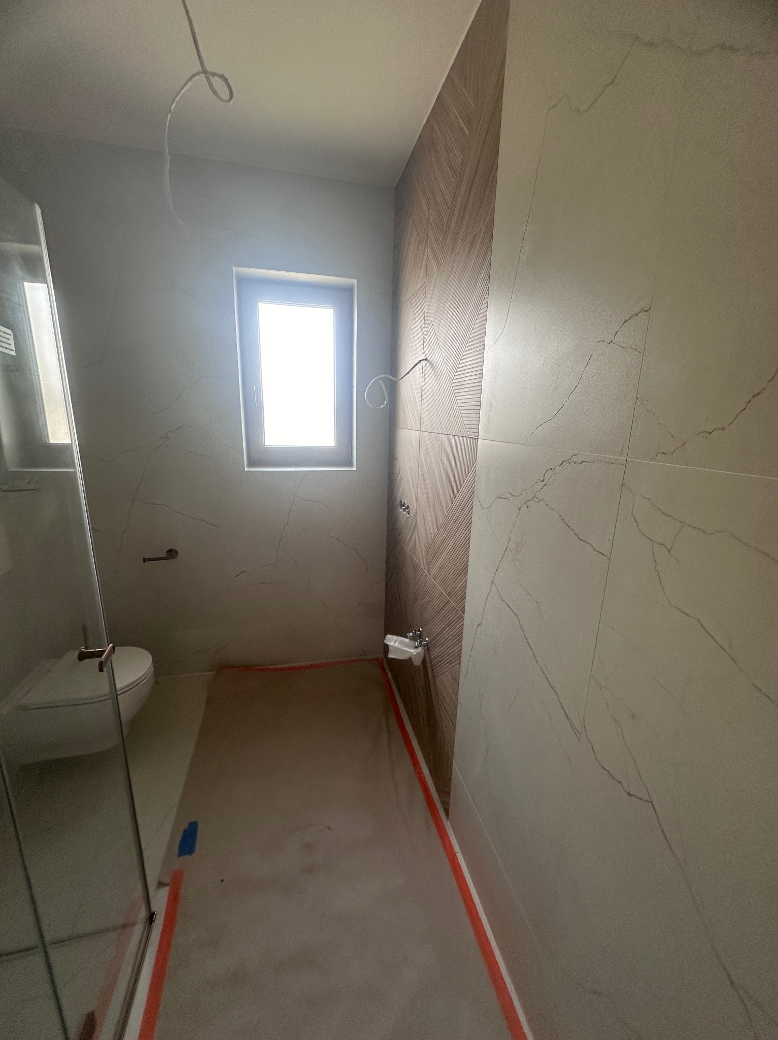 Wnętrze łazienki w trakcie remontu: widoczne płytki imitujące marmur na ścianach, częściowo ułożona podłoga i instalacja elektryczna.