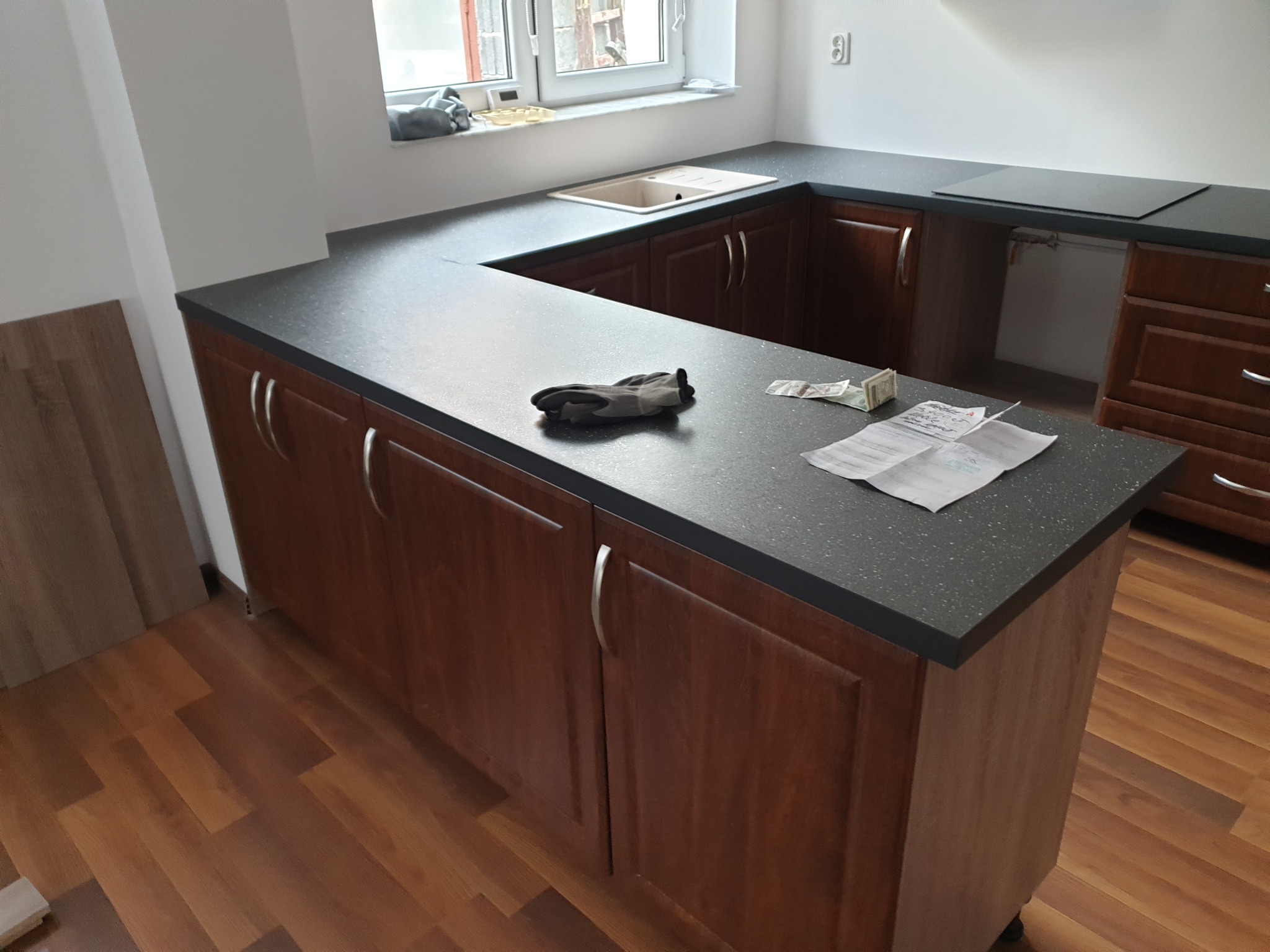Fragment nowej kuchni z drewnianymi szafkami i ciemnym blatem, na którym leżą robocze rękawice, banknoty i dokumenty, widok na okno i fragment podłogi z paneli.
