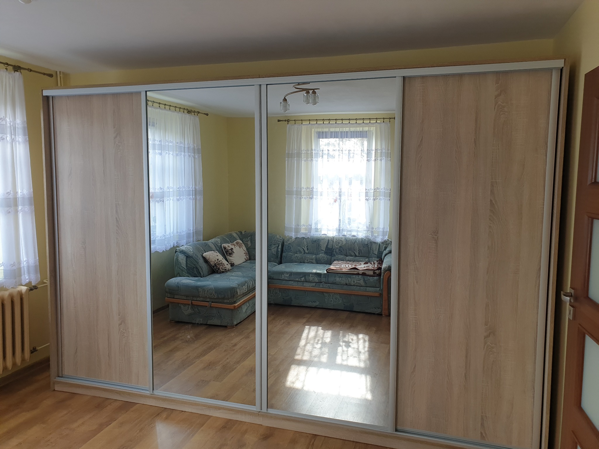 Duża szafa przesuwna z lustrzanymi drzwiami, odbijająca wnętrze pokoju z narożną sofą w kwiatowy wzór i drewnianą podłogą, oświetlona przez światło słoneczne wpadające przez firanki.